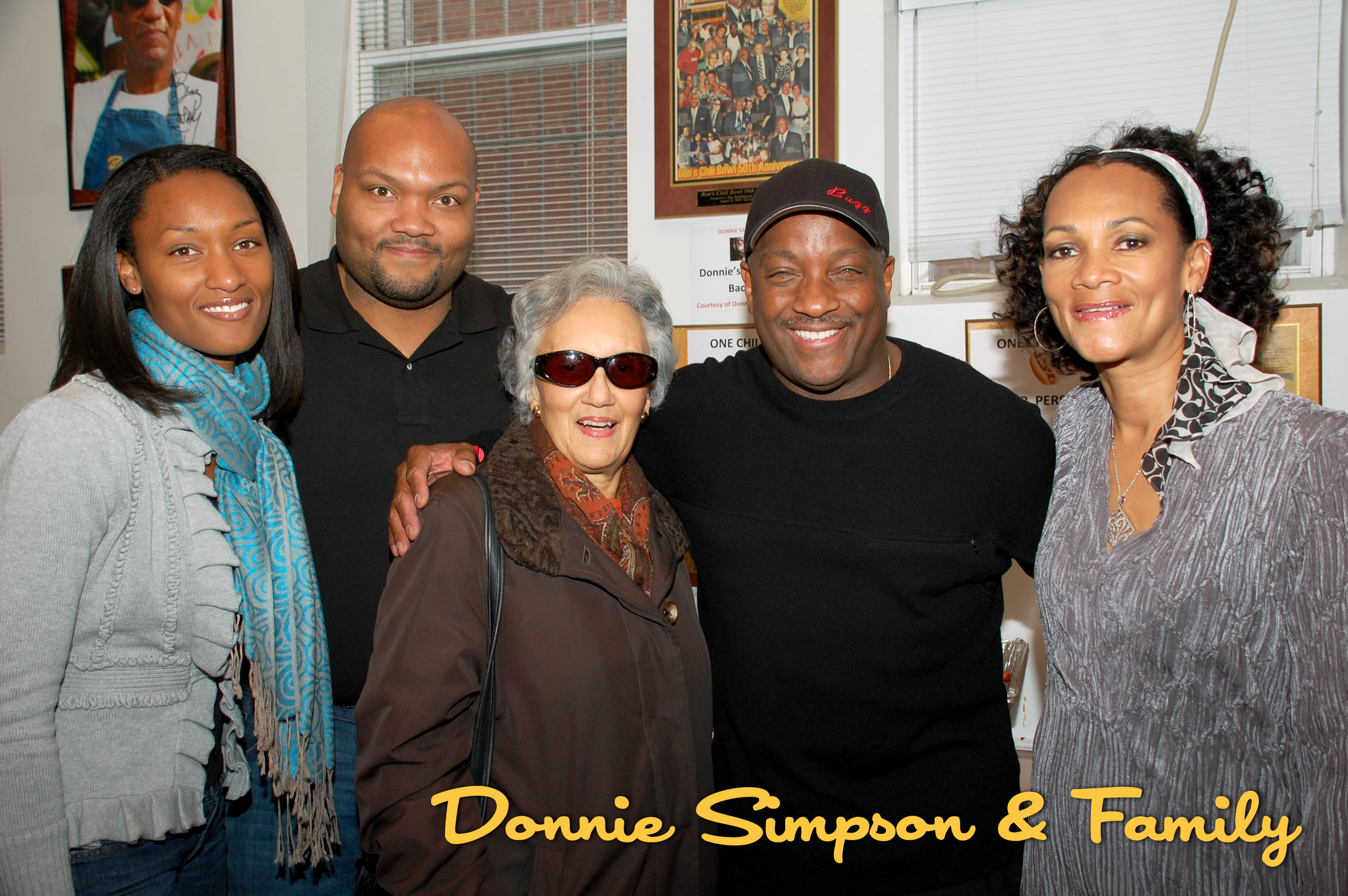 A group of six diverse people smiling indoors, with framed photos and awards on the wall behind them, captioned 'Donnie Sumpon & Family.'