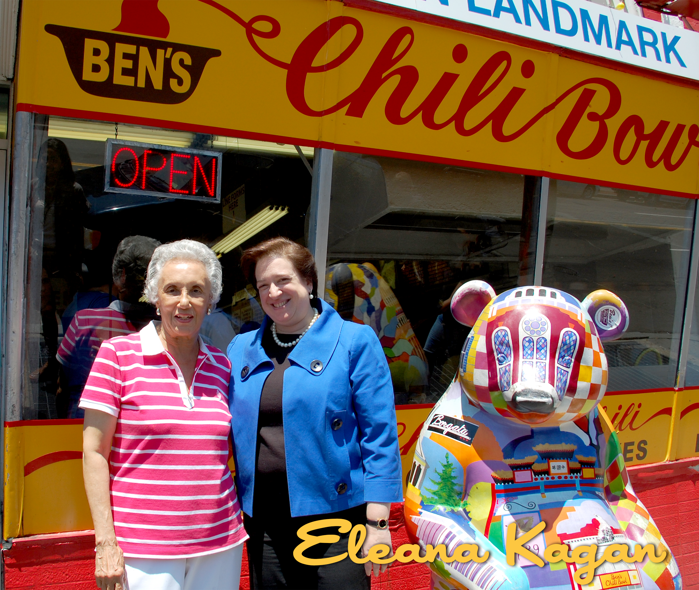 Two women standing in front of a colorful food stand with a large painted bear sculpture. The stand has a sign that reads 'Ben's Chili Bowl' with an electric 'Open' sign. One woman is wearing a pink and white striped polo shirt, and the other is wear