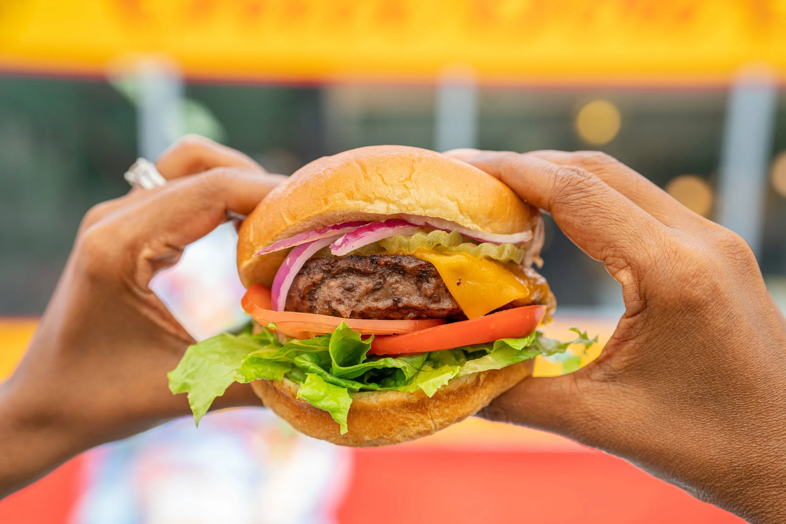 Person holding a cheeseburger with lettuce, tomato, pickles, onions, cheese, and a beef patty.