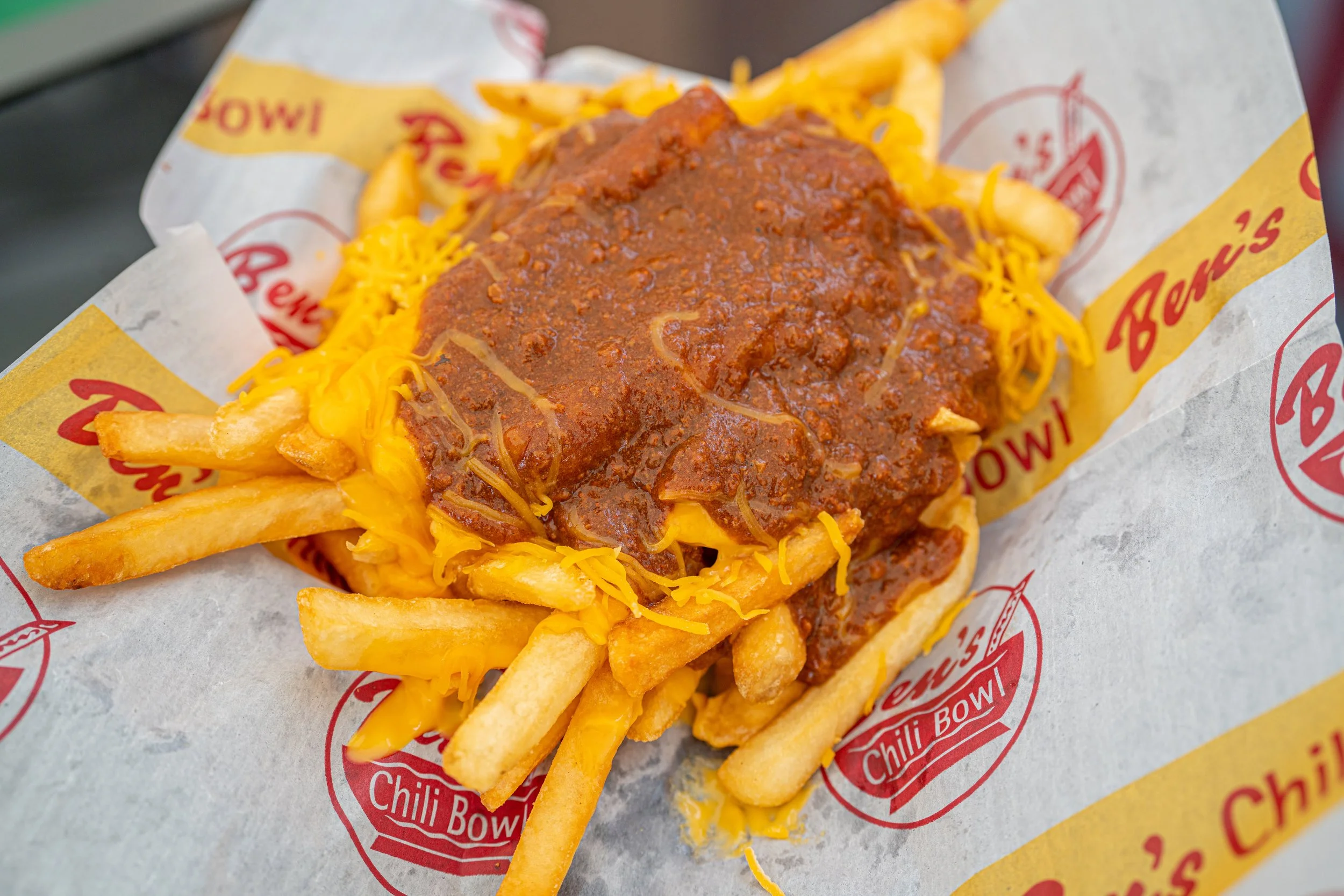 French fries topped with chili and shredded cheese in a takeout basket lined with Chili Bowl branded paper.