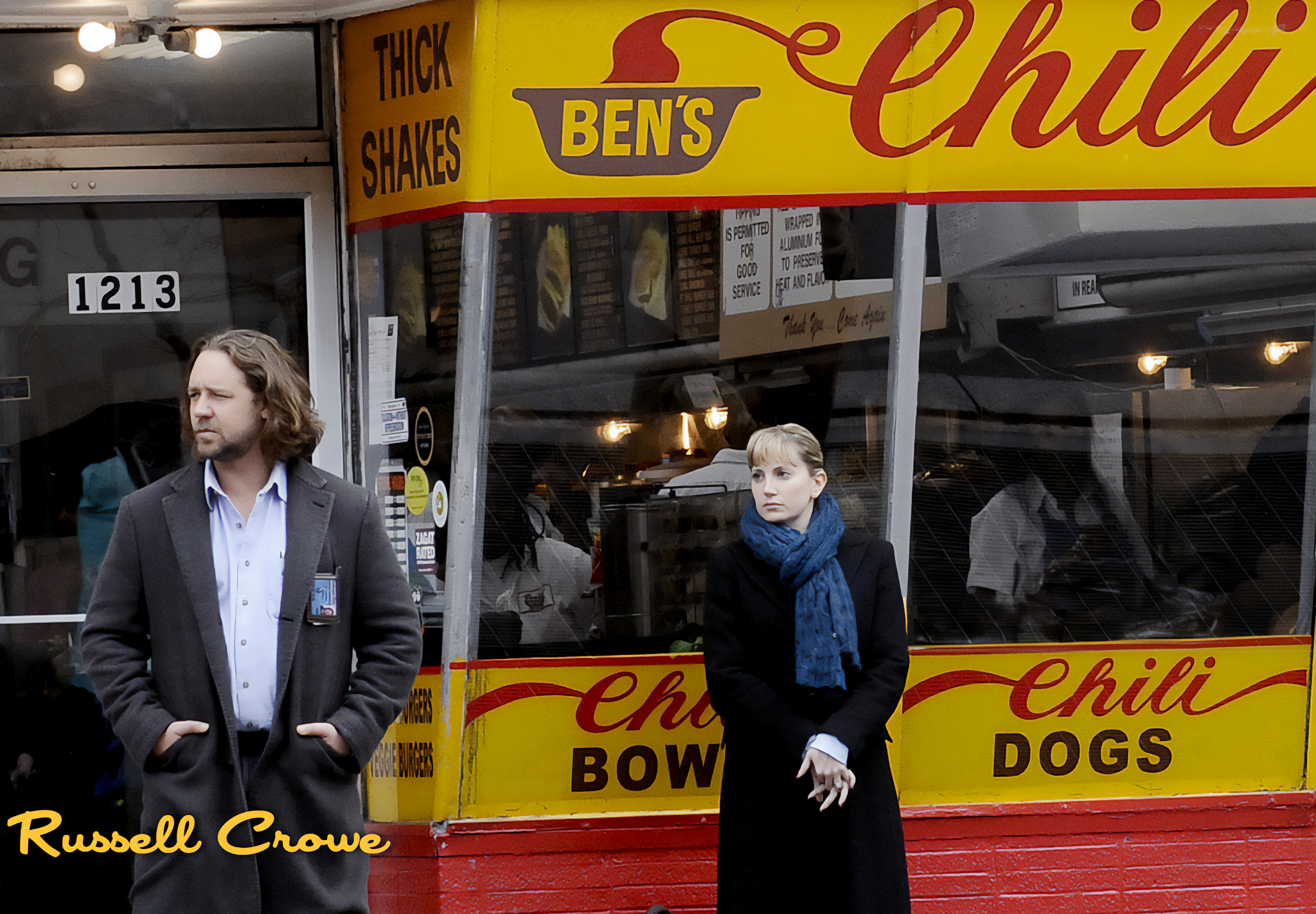 A man and a woman standing outside a fast food restaurant with a yellow and red sign that reads "Ben's Chili Dogs." The man has shoulder-length hair, wears a gray coat over a white shirt, and has his hands in his pockets. The woman has blonde hair, w