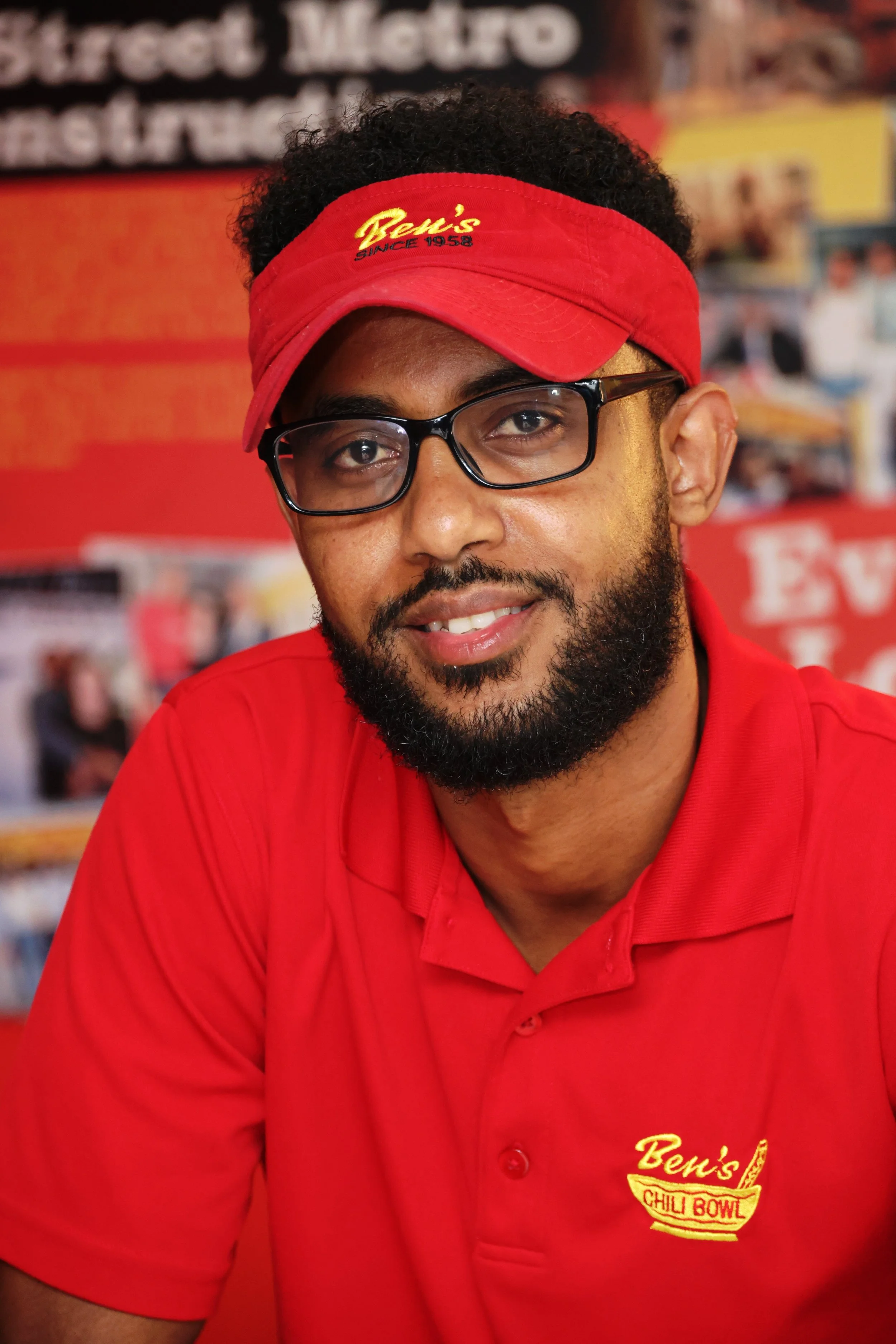 A man in red uniform with a red visor and black glasses, smiling at the camera inside Ben's Chili Bowl restaurant.