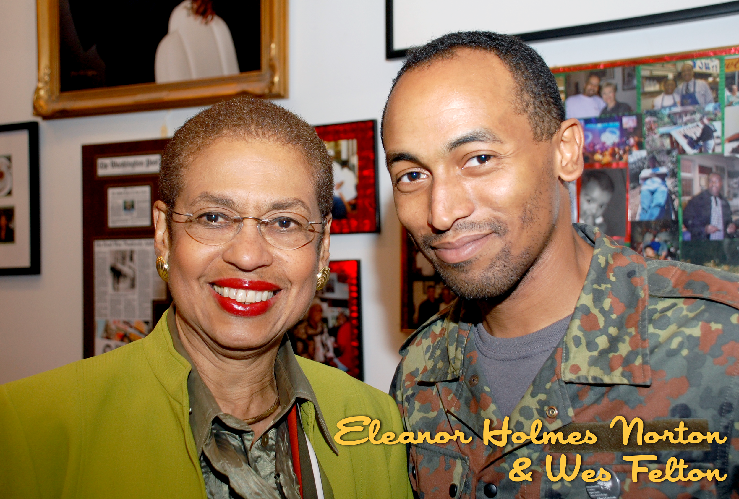Two people standing close together, smiling, with framed photos and newspaper clippings on the wall behind them. The person on the left is an older woman with short hair, glasses, red lipstick, and wearing a lime green blazer. The person on the right
