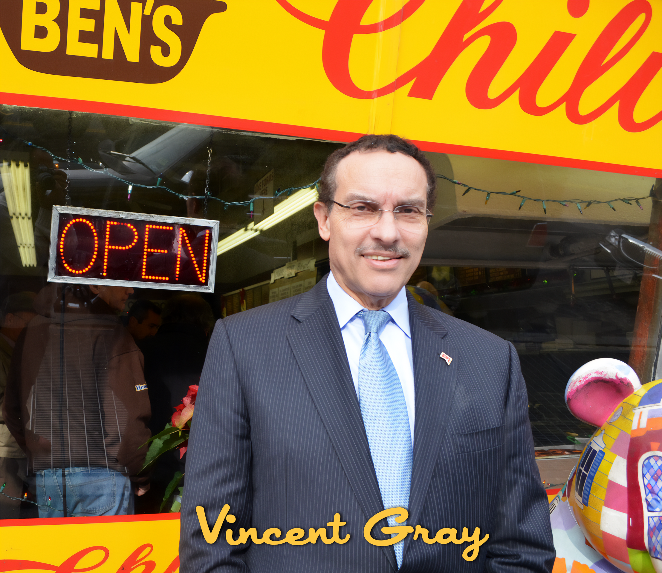 Man in a business suit standing outside a storefront with a bright yellow and red sign that reads 'Ben's Chul'. The storefront has a neon 'Open' sign and a window showing people inside. The man has short dark hair, glasses, and a light blue tie, and 