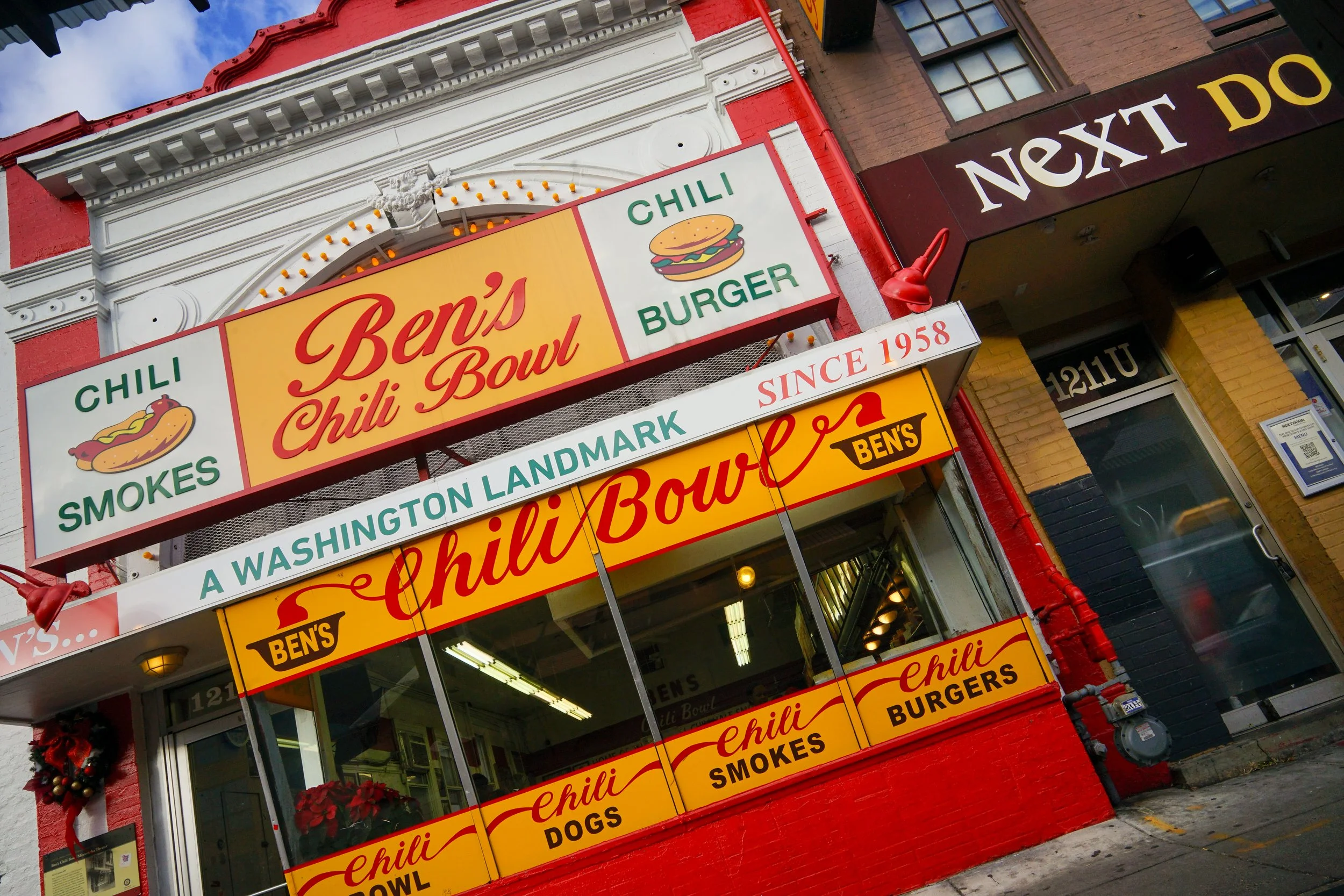 Exterior view of Ben's Chili Bowl in Washington, D.C., featuring yellow and red signage advertising chili dogs, chili smokes, chili burgers, and that the establishment has been serving since 1958 with decorative holiday wreaths on the storefront.
