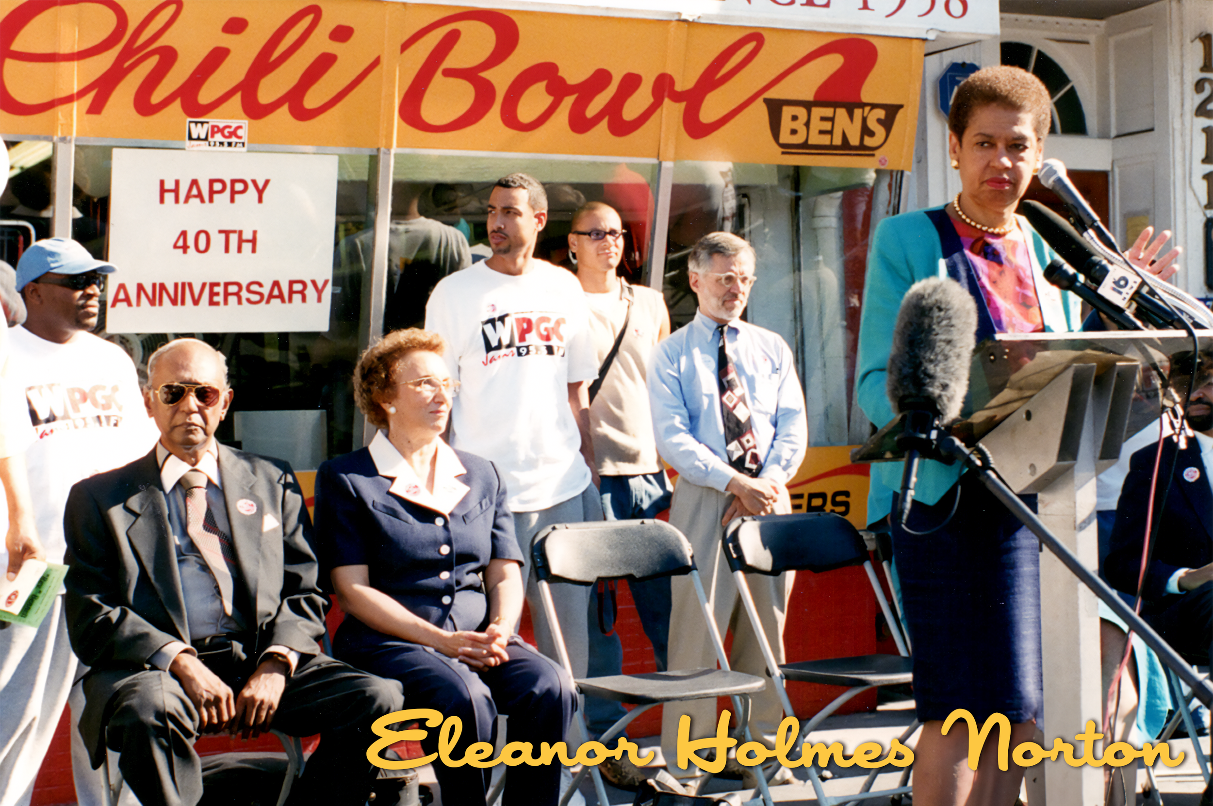 Eleanor Holmes Norton speaking at a podium during a celebration event, with a group of people standing and sitting behind her, in front of a storefront with a sign that reads 'Happy 40th Anniversary' and 'Chili Bowl'.