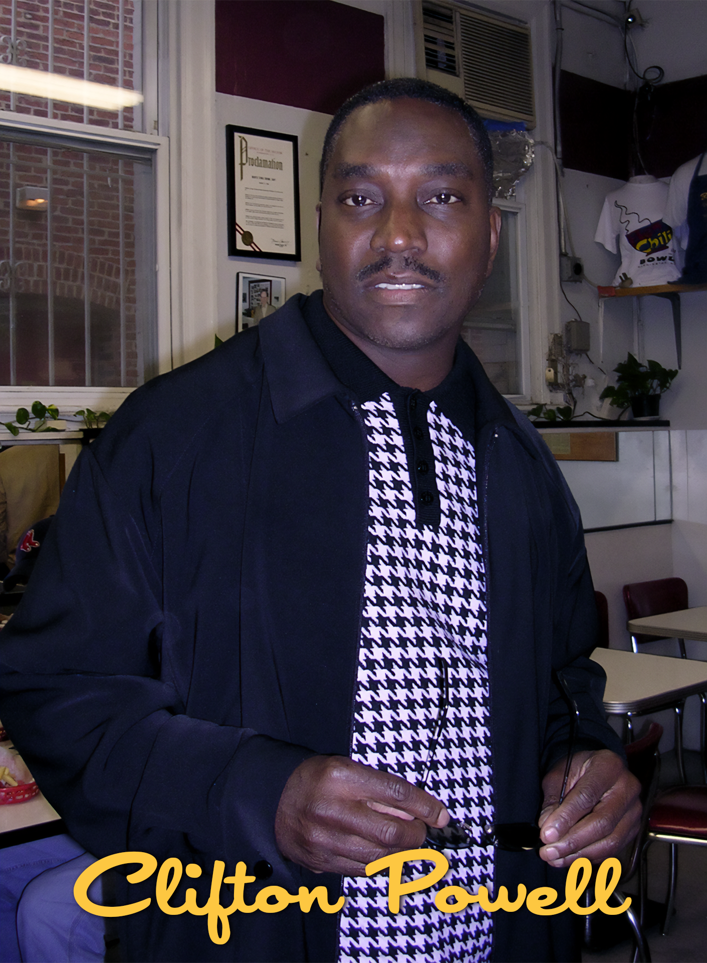 A man wearing a black jacket and a black-and-white houndstooth shirt holding sunglasses inside a room with pictures and certificates on the wall.