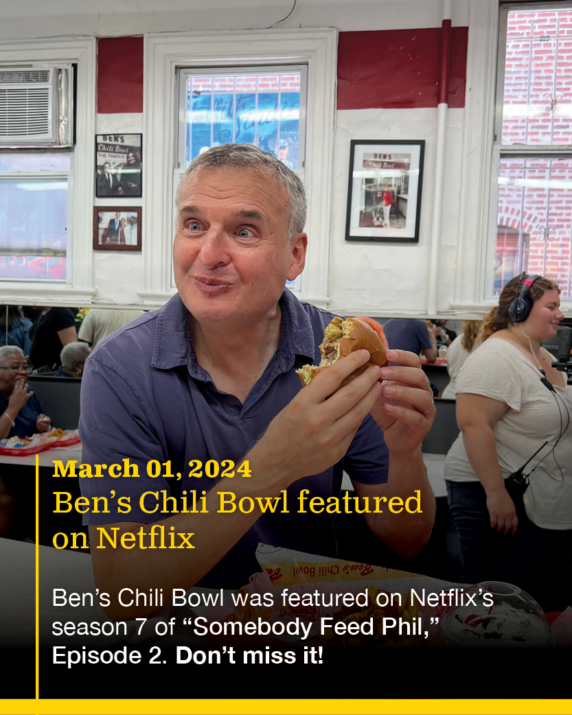 A man sitting in a restaurant holding a burger, with people and framed photos on the wall behind him, and a woman wearing headphones in the background.