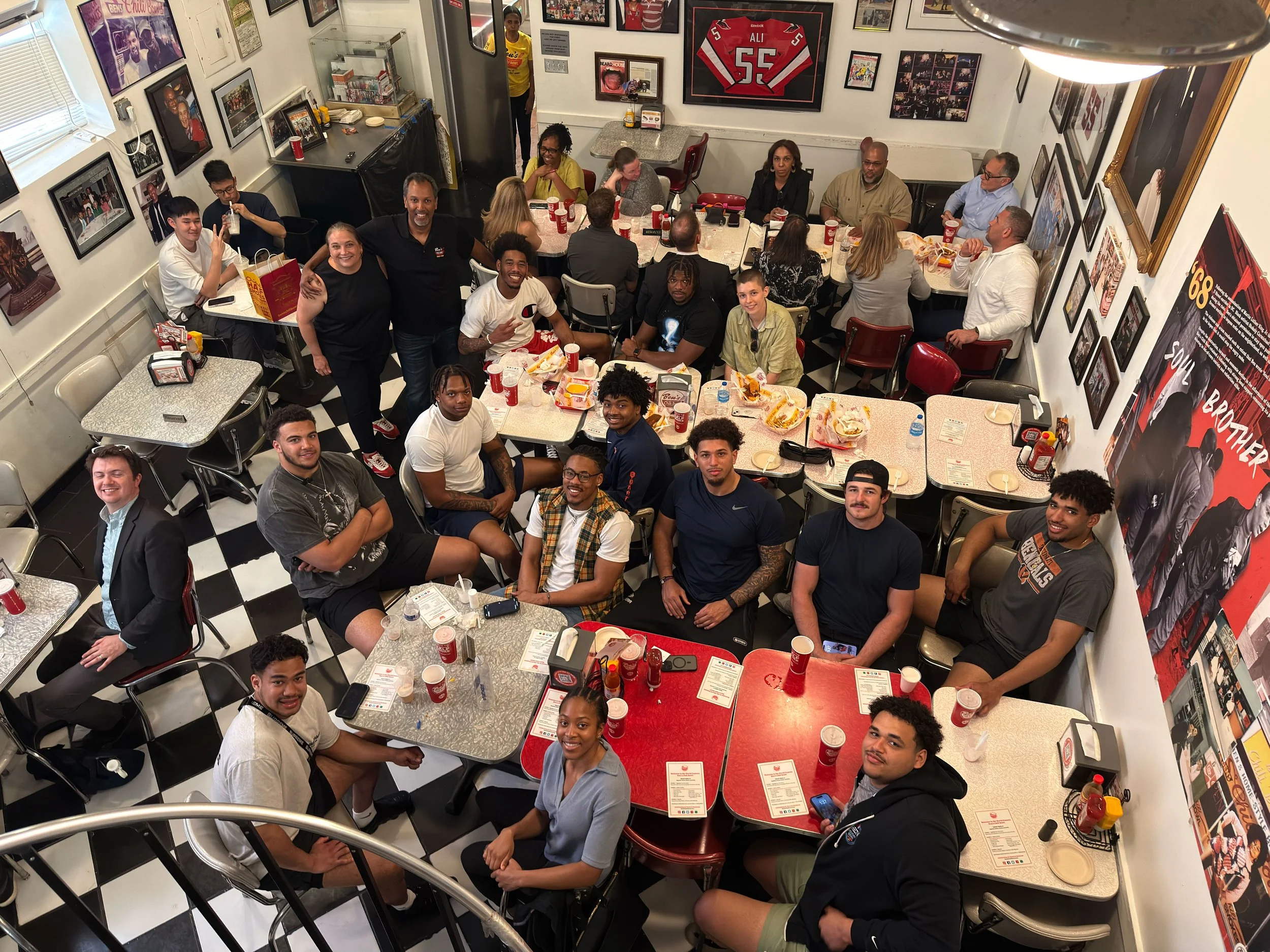 A group of young people and some adults gathered inside a restaurant, posing for a photo. They sit and stand around tables with food and drinks, with walls decorated with sports memorabilia and framed photos.