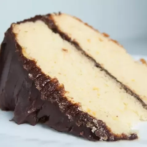 A slice of vanilla cake with chocolate frosting and a chocolate coating on the outside