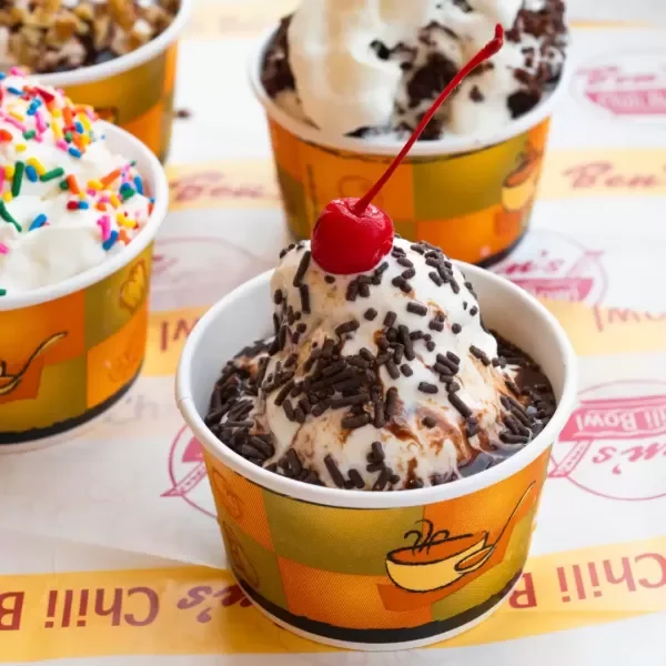 A cup of ice cream topped with sprinkles and chocolate syrup, garnished with a cherry, placed on a paper fast food tray, with more cups of ice cream in the background.