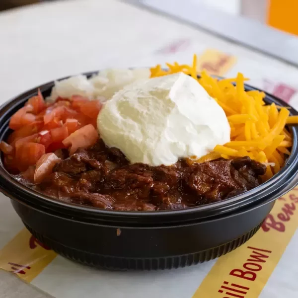 A black bowl of chili topped with shredded cheddar cheese, sour cream, diced tomatoes, and a baked potato.