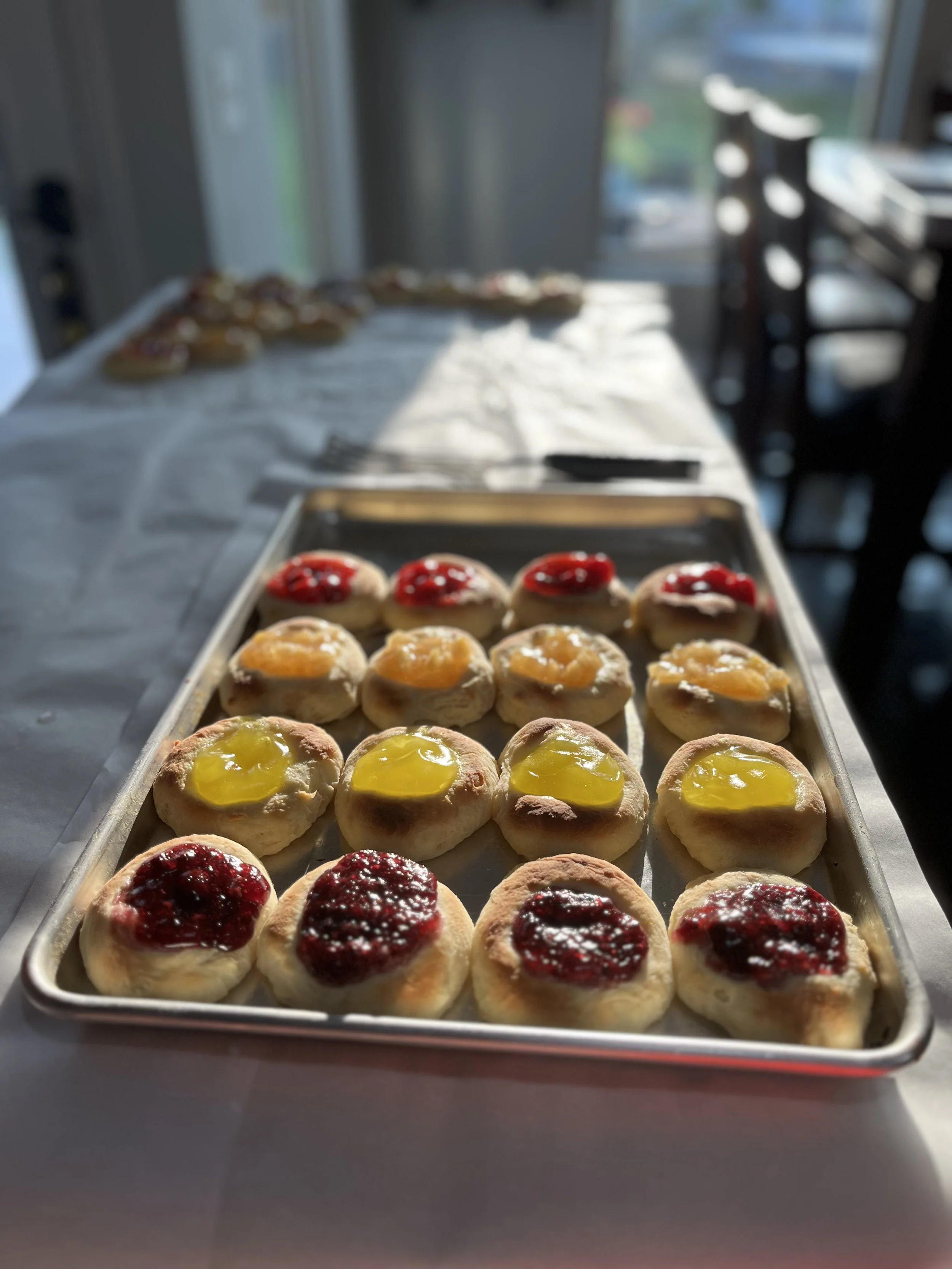 Tray of kolaches with various fruit preserves including raspberry, lemon, and cherry, on a kitchen table with sunlight.