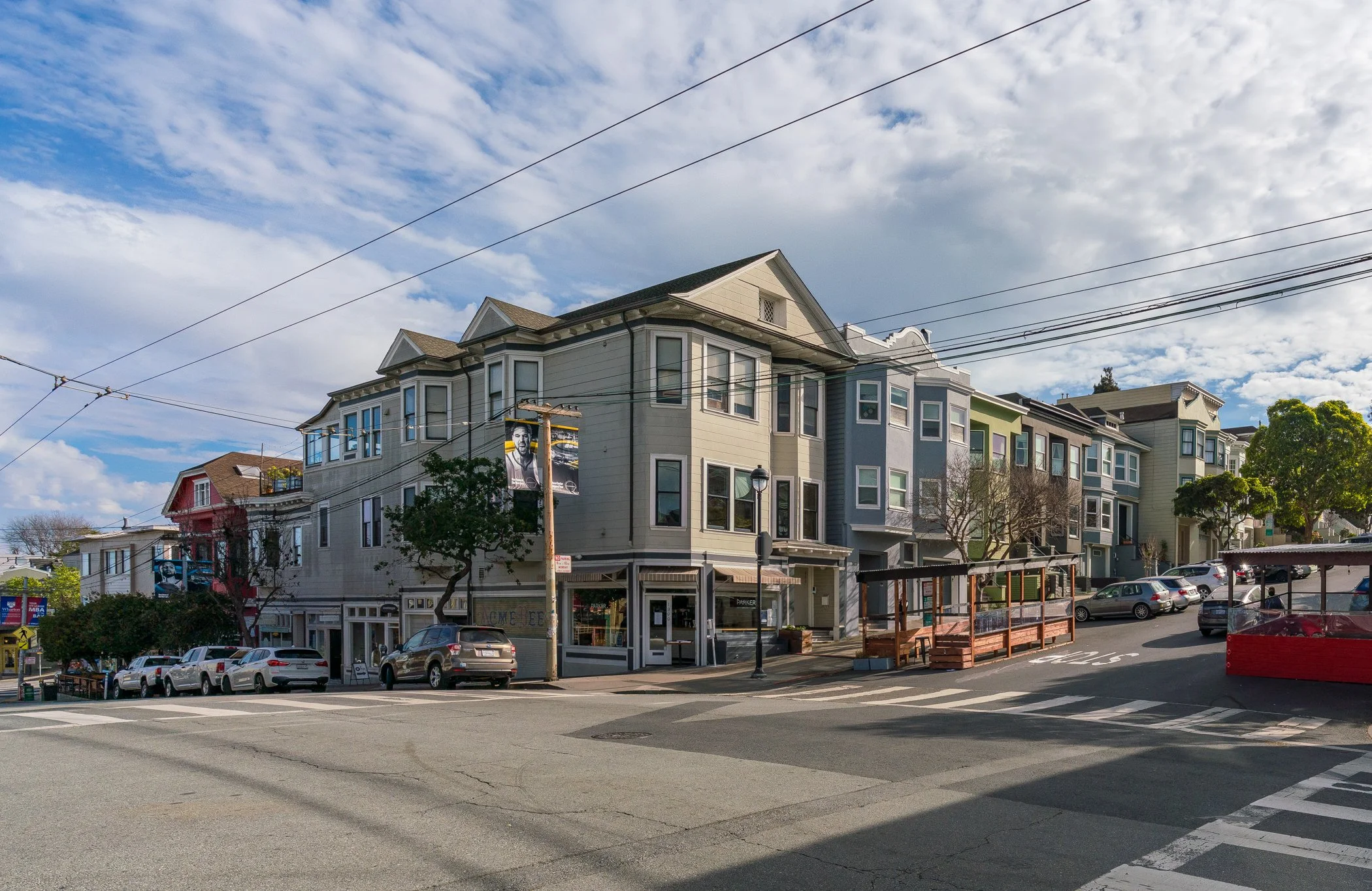 A street scene with colorful multi-story houses on a sloped hill, parked cars, power lines, and a partly cloudy sky.