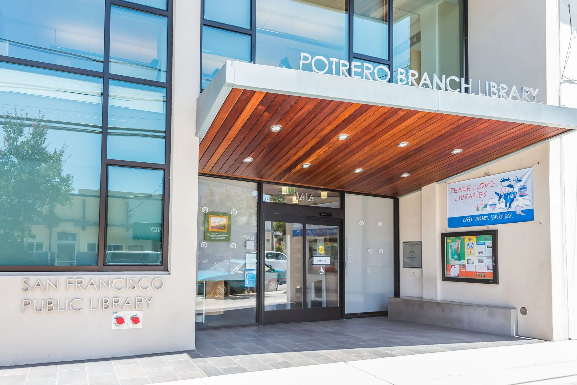 Entrance of the Potrero Branch Library in San Francisco with a wooden ceiling over the doorway, glass windows, and a banner that reads 'Peace, Love, Libraries.'