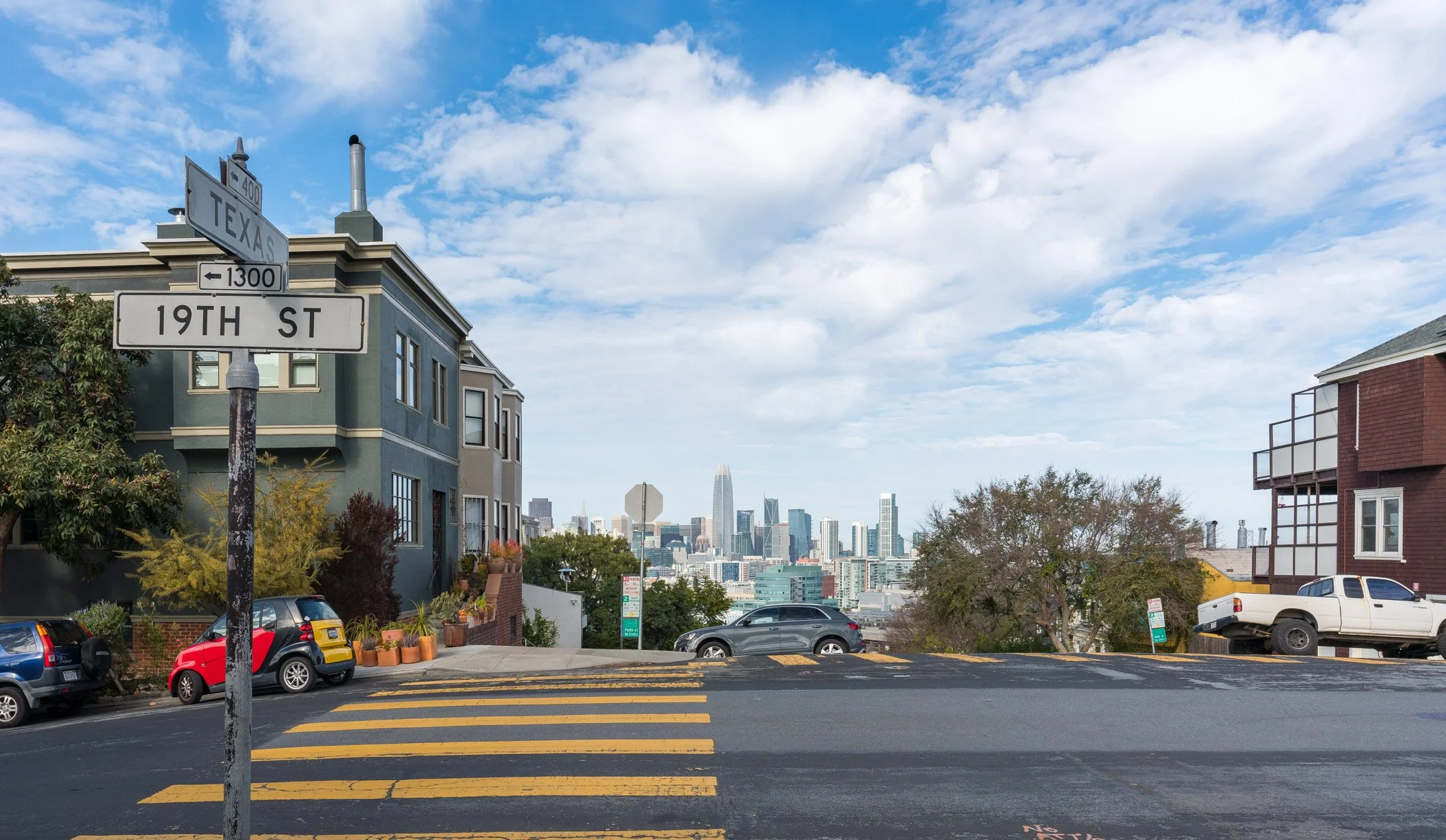 Street view at the intersection of 19th Street and Texas with a city skyline in the background, parked cars, and residential buildings.