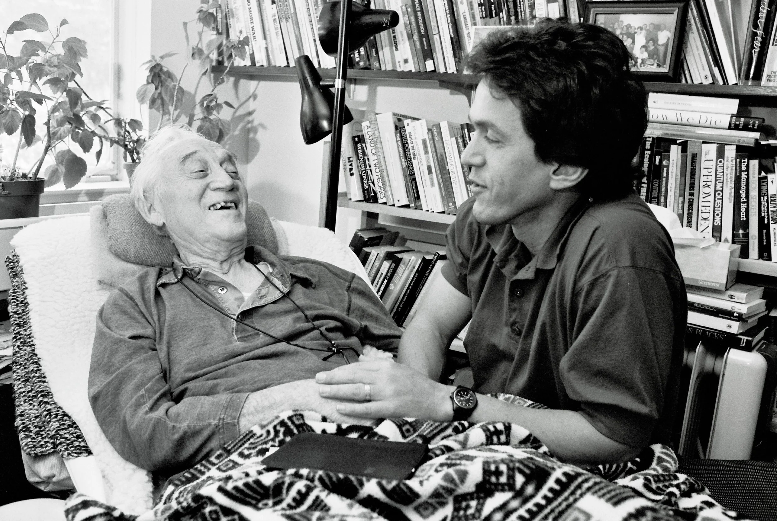 Mitch Albom holding the hand of his former professor Morrie Schwartz as they sit together in Morrie’s home, surrounded by books.