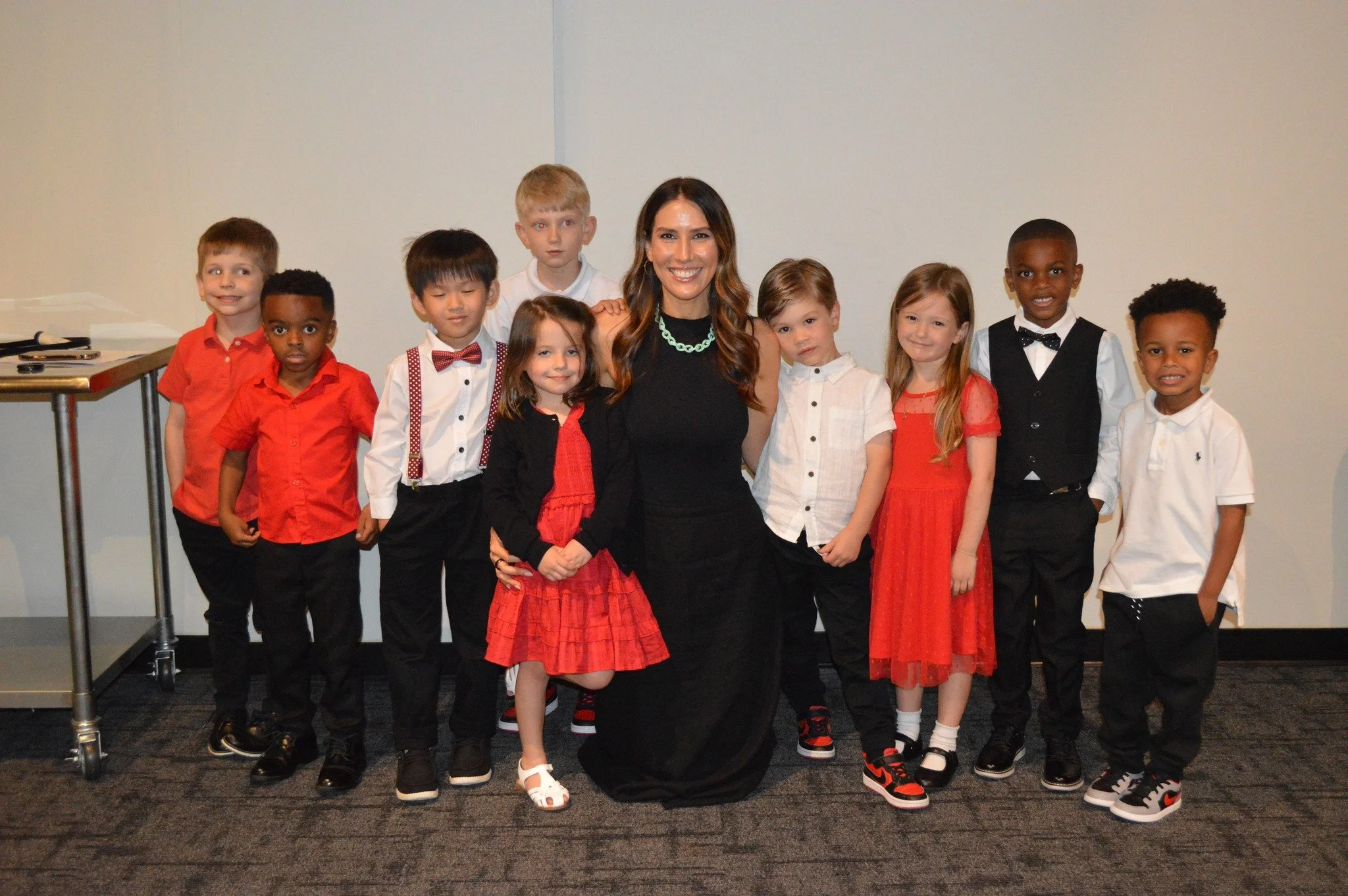 Group of children dressed in semi-formal clothing standing with a woman in a black dress, all smiling for a photo against a plain background.