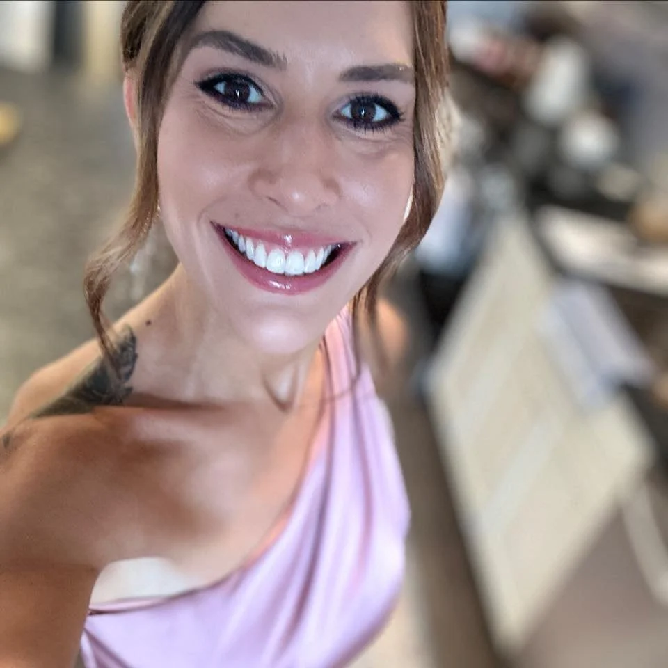 A woman smiling with white teeth in a pink top, with blurred background.