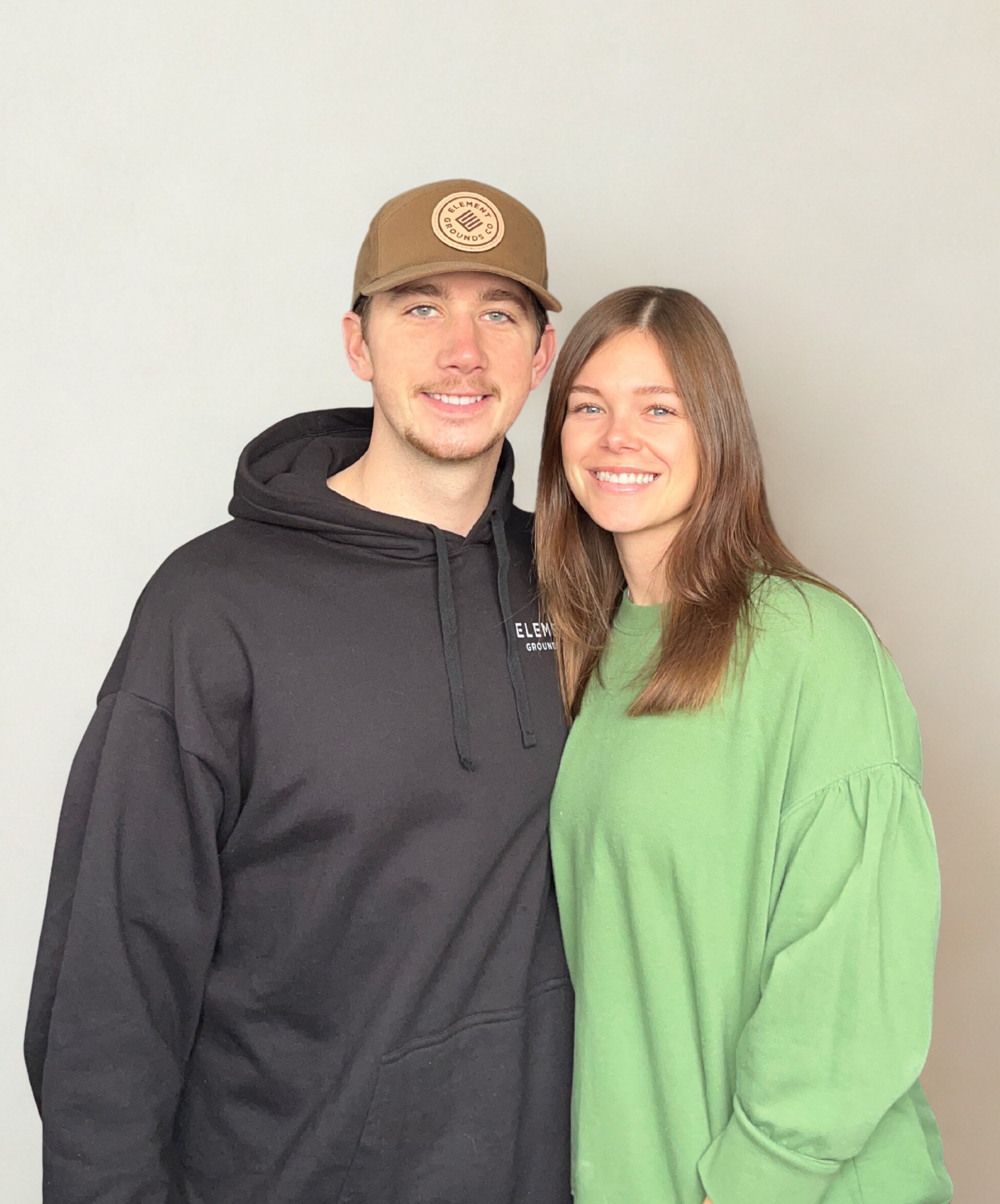 A young man and woman smiling, standing close together against a plain beige background. The man wears a brown cap and a black hoodie, and the woman wears a bright green sweatshirt.
