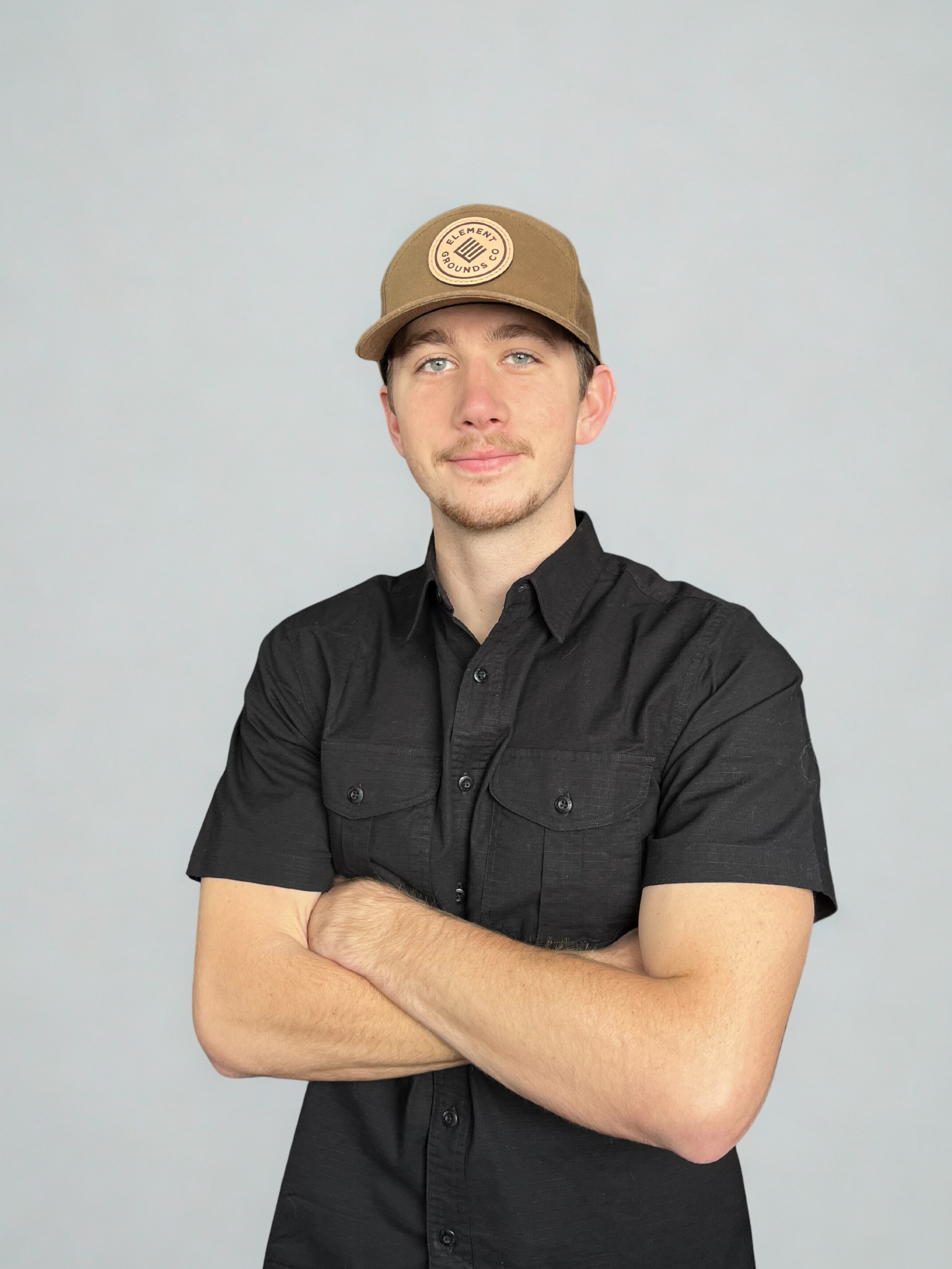 A young man with light skin, light brown hair, and blue eyes wearing a khaki baseball cap with a circular logo that says 'Element Grounds Co.' and a black short-sleeved button-up shirt, standing with arms crossed against a plain light gray background.