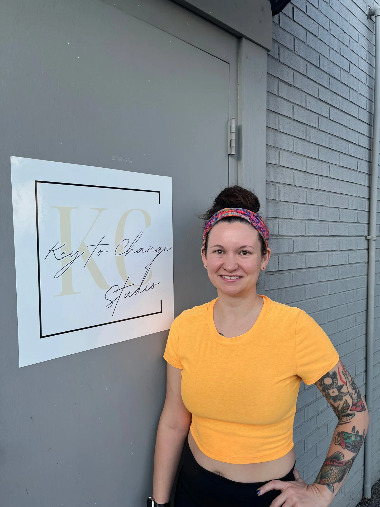 Pilates instructor, Sara Fisher, is smiling and standing next to the Key to Change Studio door. She's wearing an orange shirt with a colorful headband.