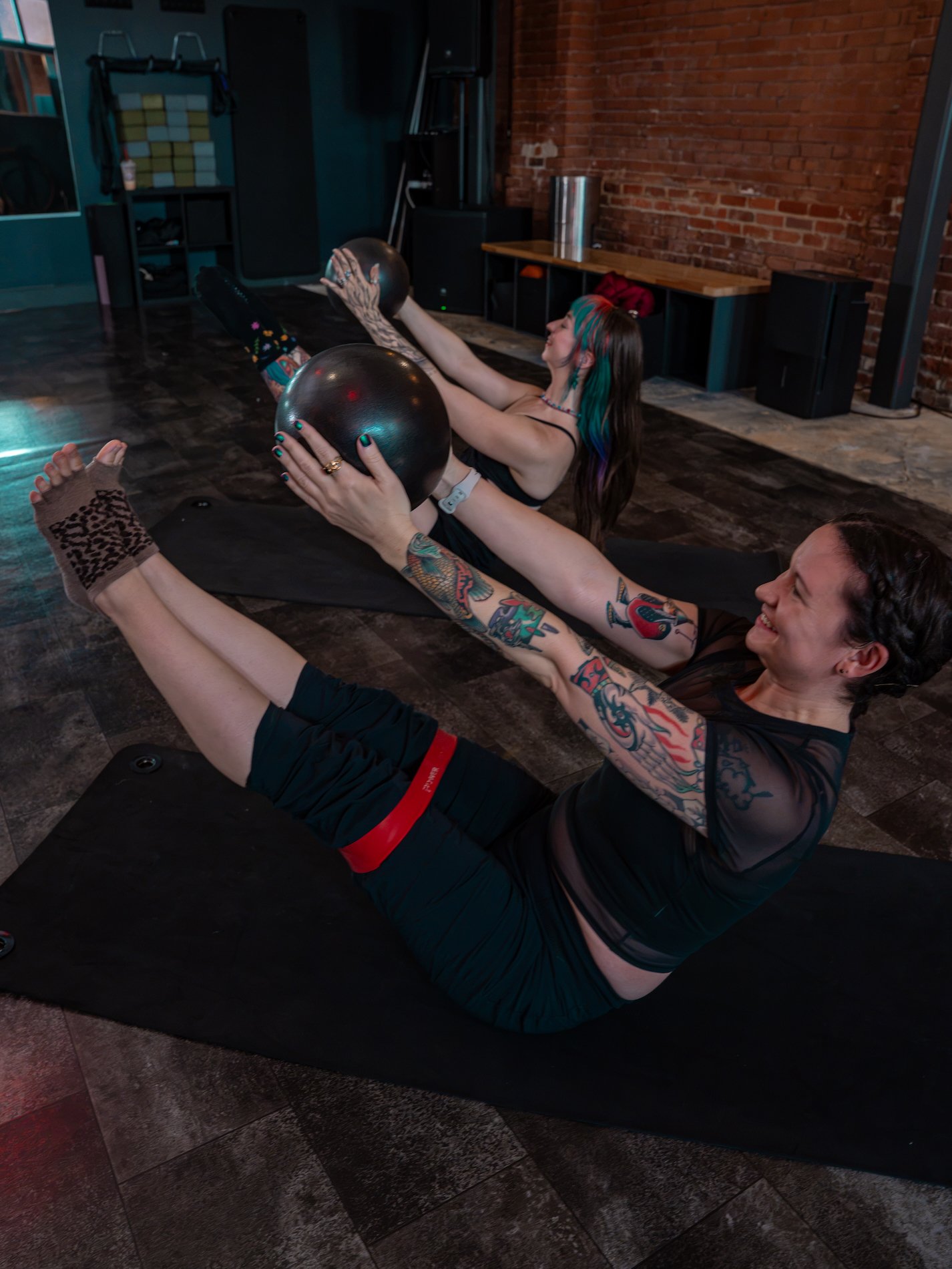 Two smiling women with tattoos and colorful hair doing mat Pilates side by side in a fitness studio, holding a Pilates ball and using resistance bands around their thighs