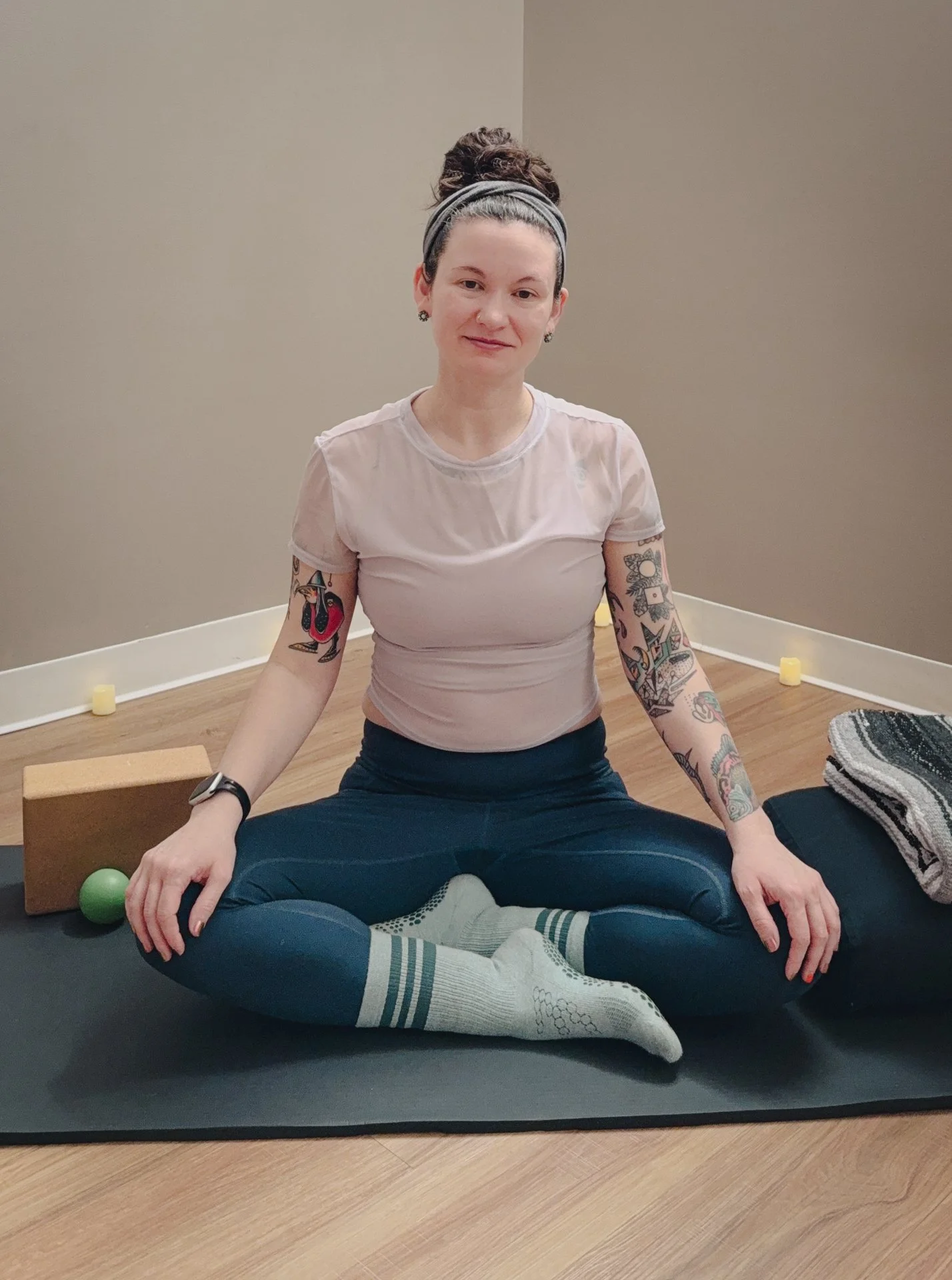 Photo of Sara sitting cross-legged on a yoga mat and smiling, resting her hands on her knees. Yoga props can be seen in the background.
