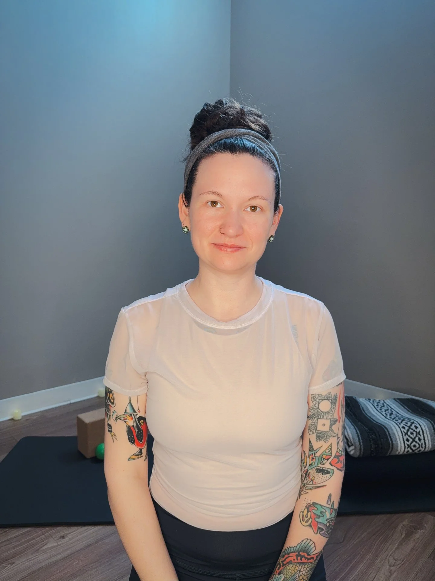 Portrait of Sara smiling with hair pulled up in a large top bun, wearing a headband. Yoga props can be seen in the background behind her.