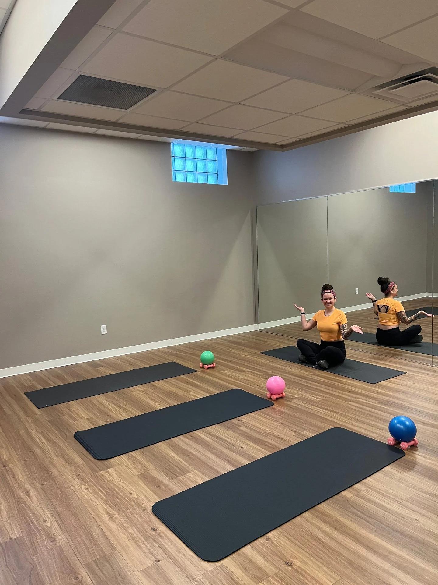 Pilates instructor, Sara Fisher, is sitting cross-legged on a black exercise mat in a studio fitness room with Pilates mats set up in front of her. There is a mirrored wall behind her.