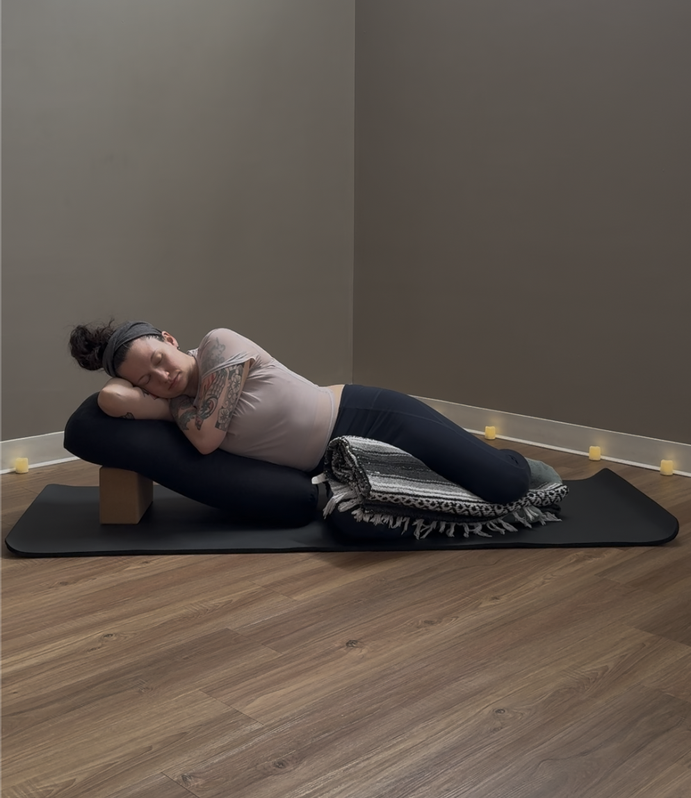 Photo of Sara doing a relaxing, side-lying restorative yoga pose. She's lying on her right sight supported by a bolster with her arms tucked comfortably under her head, using a blanket as padding between her bent knees.