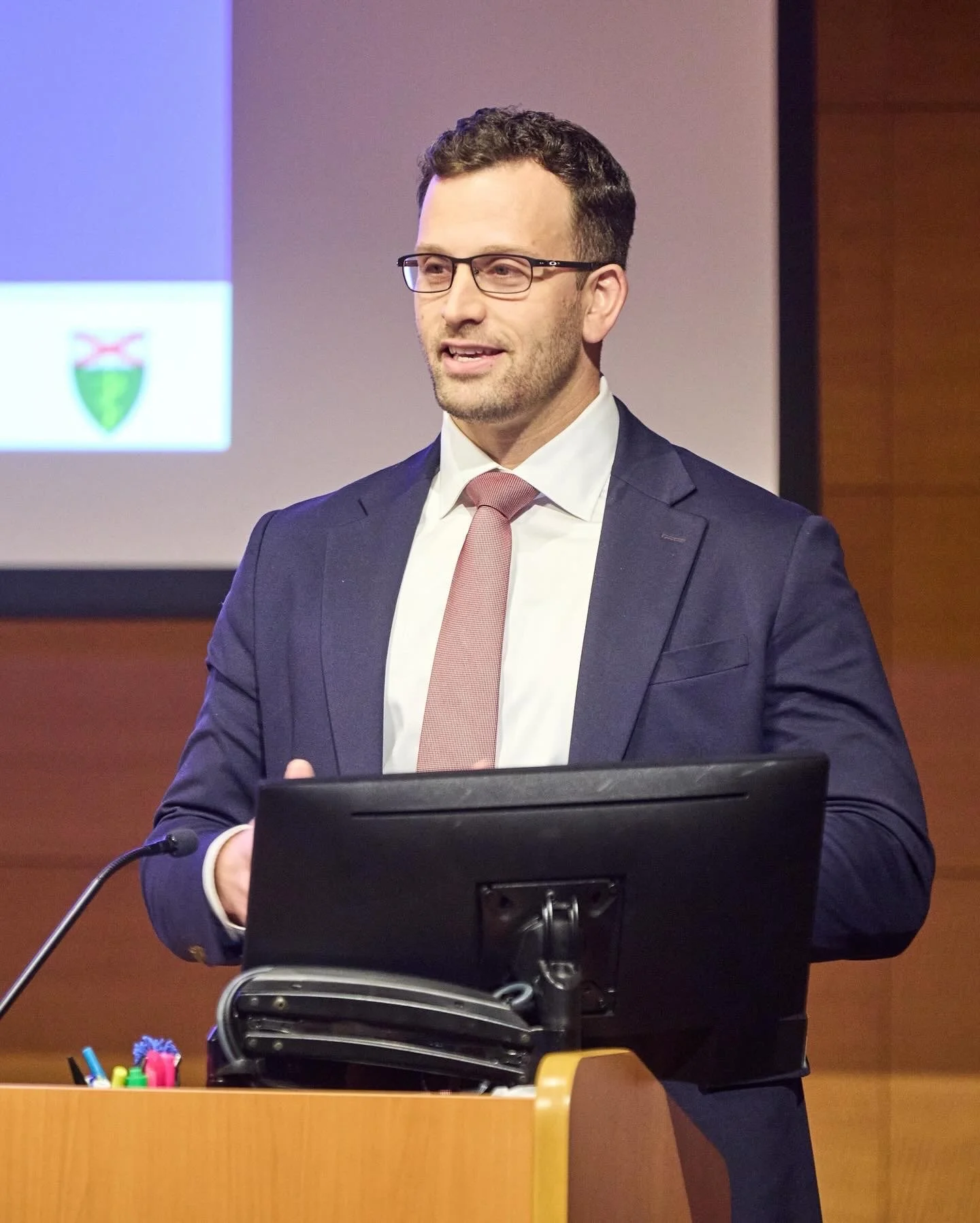 A man wearing glasses, a navy suit, white shirt, and pink tie standing at a podium, speaking in a conference room with a presentation projected on the wall behind him.