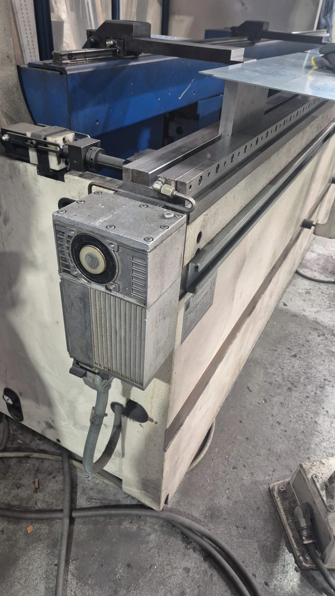 Máquina industrial con componentes metálicos, parte superior con una barra y una herramienta, y un motor en la esquina inferior izquierda, en un entorno de taller o fábrica.