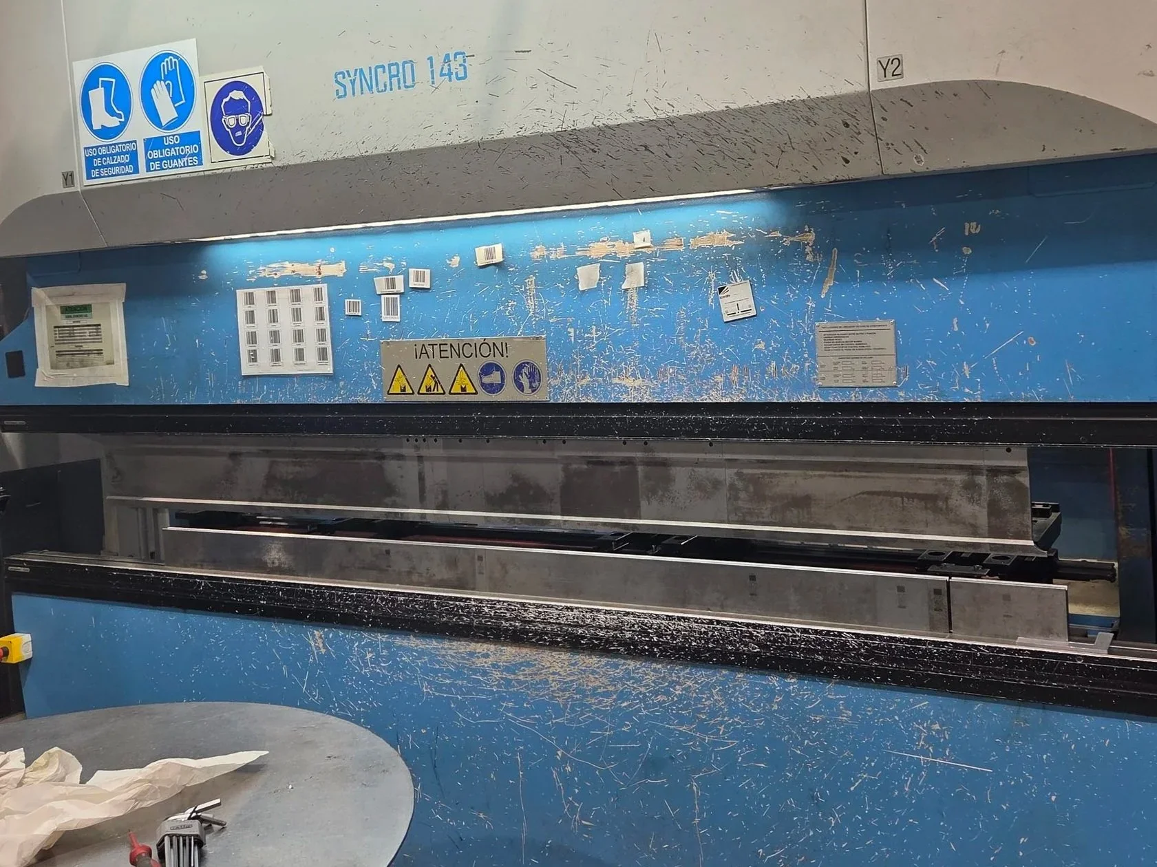 Vista de una cabina de inspección en un aeropuerto o entidad de seguridad, con signos de advertencia y procedimientos obligatorios como uso de calzado y guantes, y señales de seguridad en la parte superior. La cabina está sucia y con herramientas sobre una mesa en primer plano.