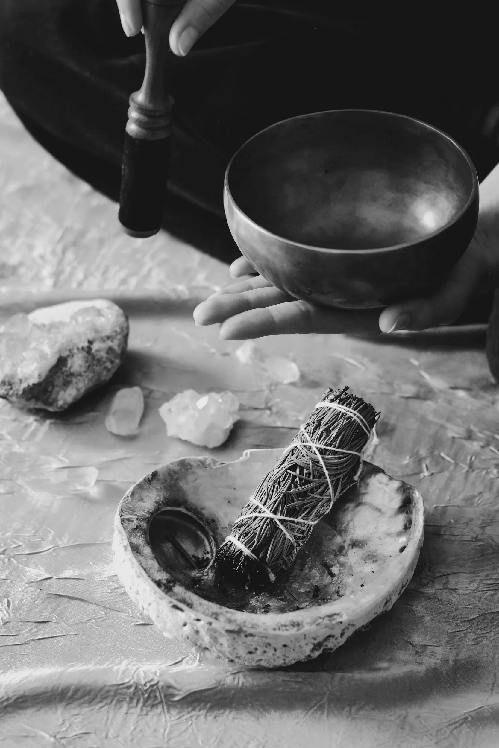The image depicts a hand holding a sound bowl, as well as tied bundles of dried herbs, and crystals on the ground in front.