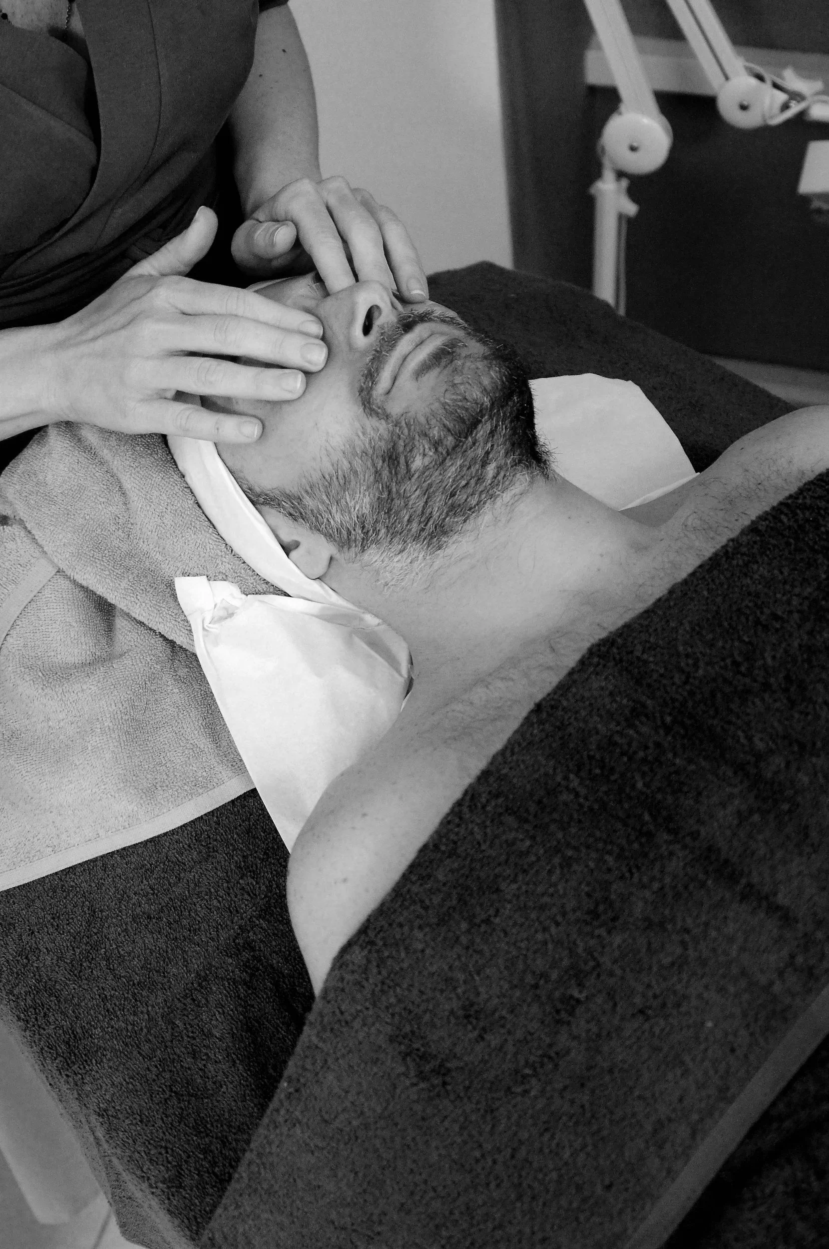 A man lays on an esthetician's table with his eyes closed and a blanket over him. The esthetician's hands gently rest on his eyes. The photo is from the chest up.