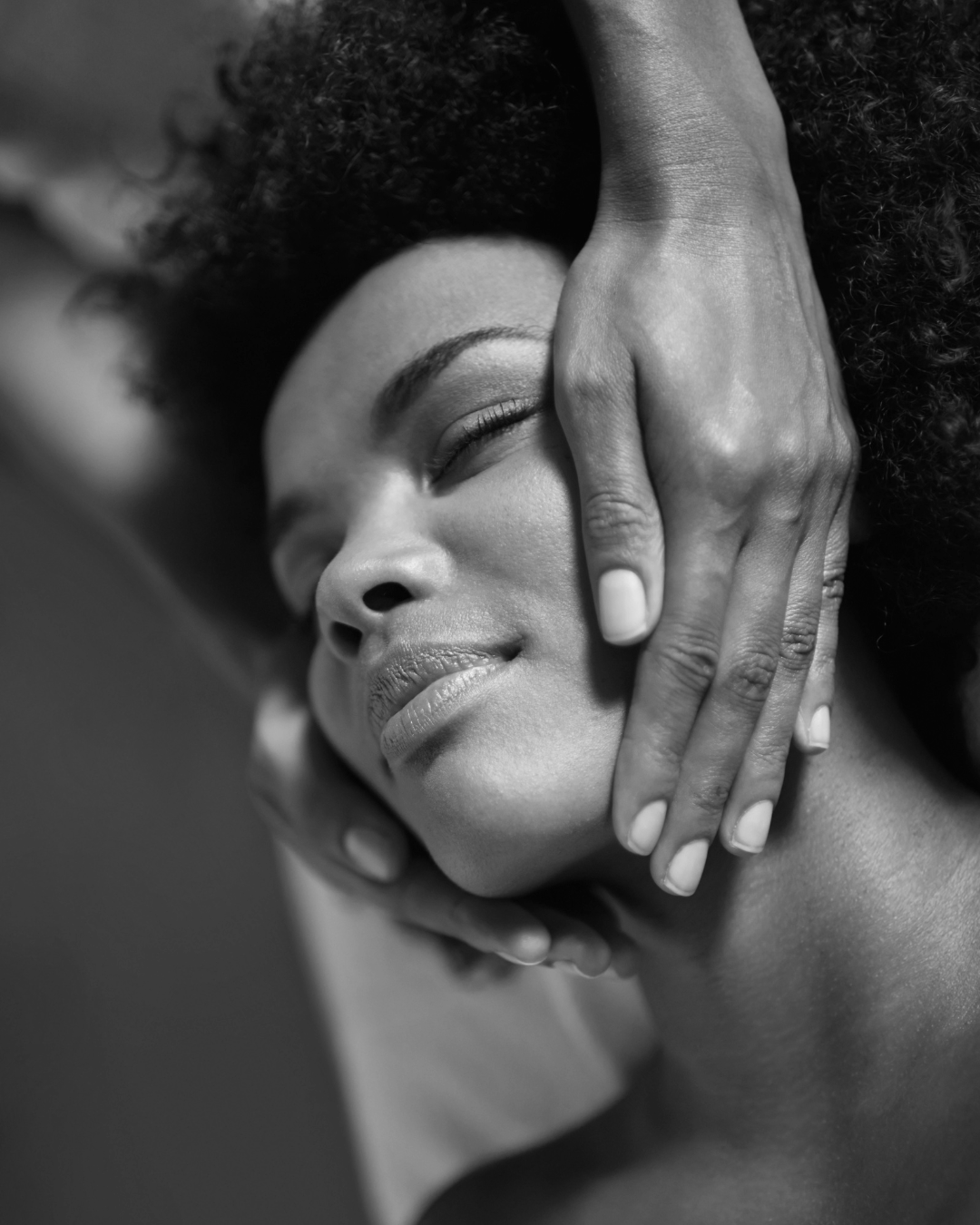 A Black woman is laying down with her eyes closed, while hands gently cup her face. The photo is from the neck up.