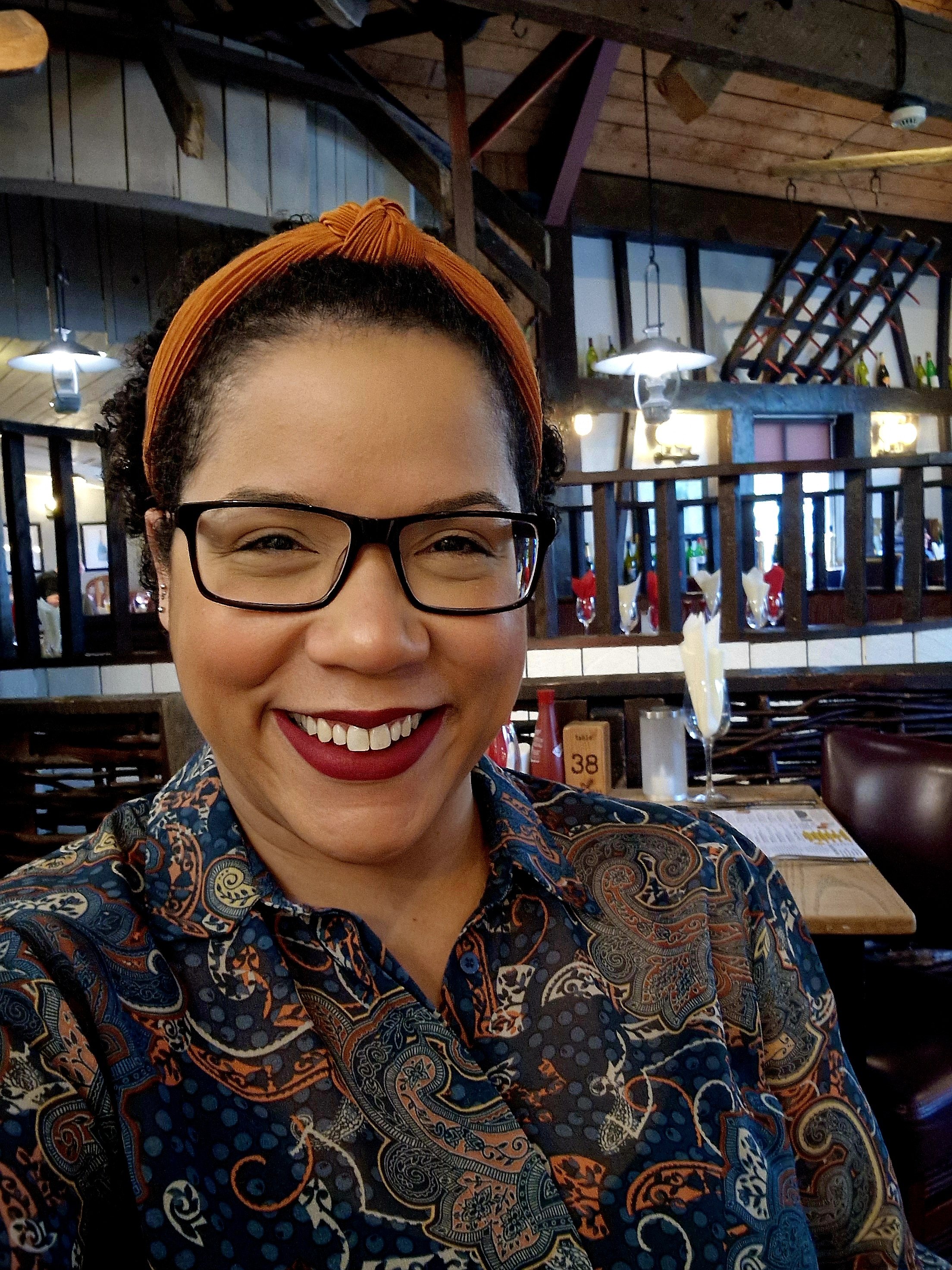 A woman with curly hair, glasses, and red lipstick smiling in a restaurant with wooden decor and table settings in the background.