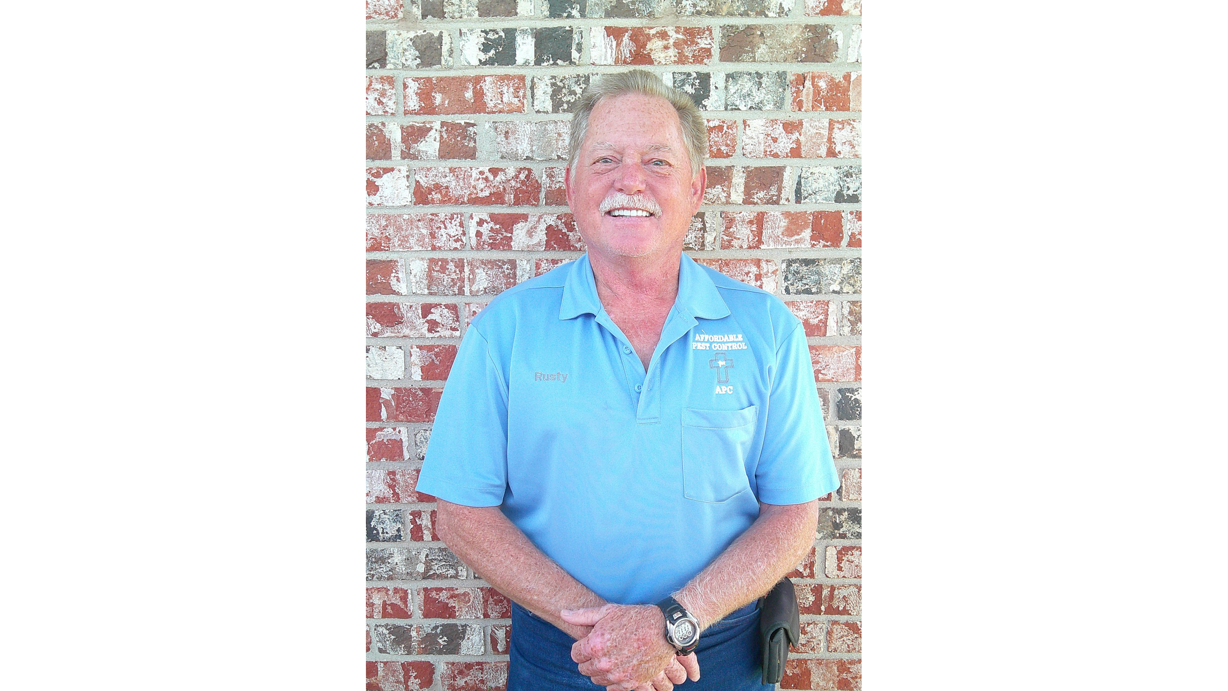 A person wearing a beekeeping suit and veil, smiling, standing outside near a building with a high voltage sign.