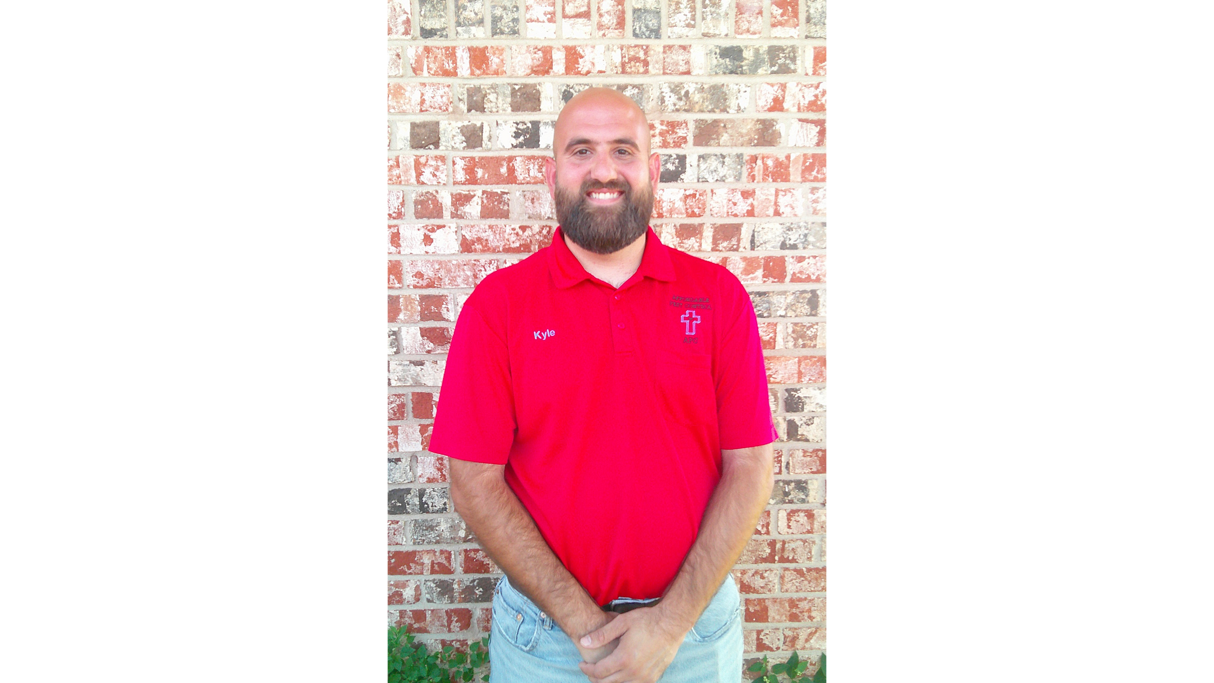 Man with a beard wearing a red polo shirt and jeans standing in front of a blue pickup truck with religious and service vehicle stickers.