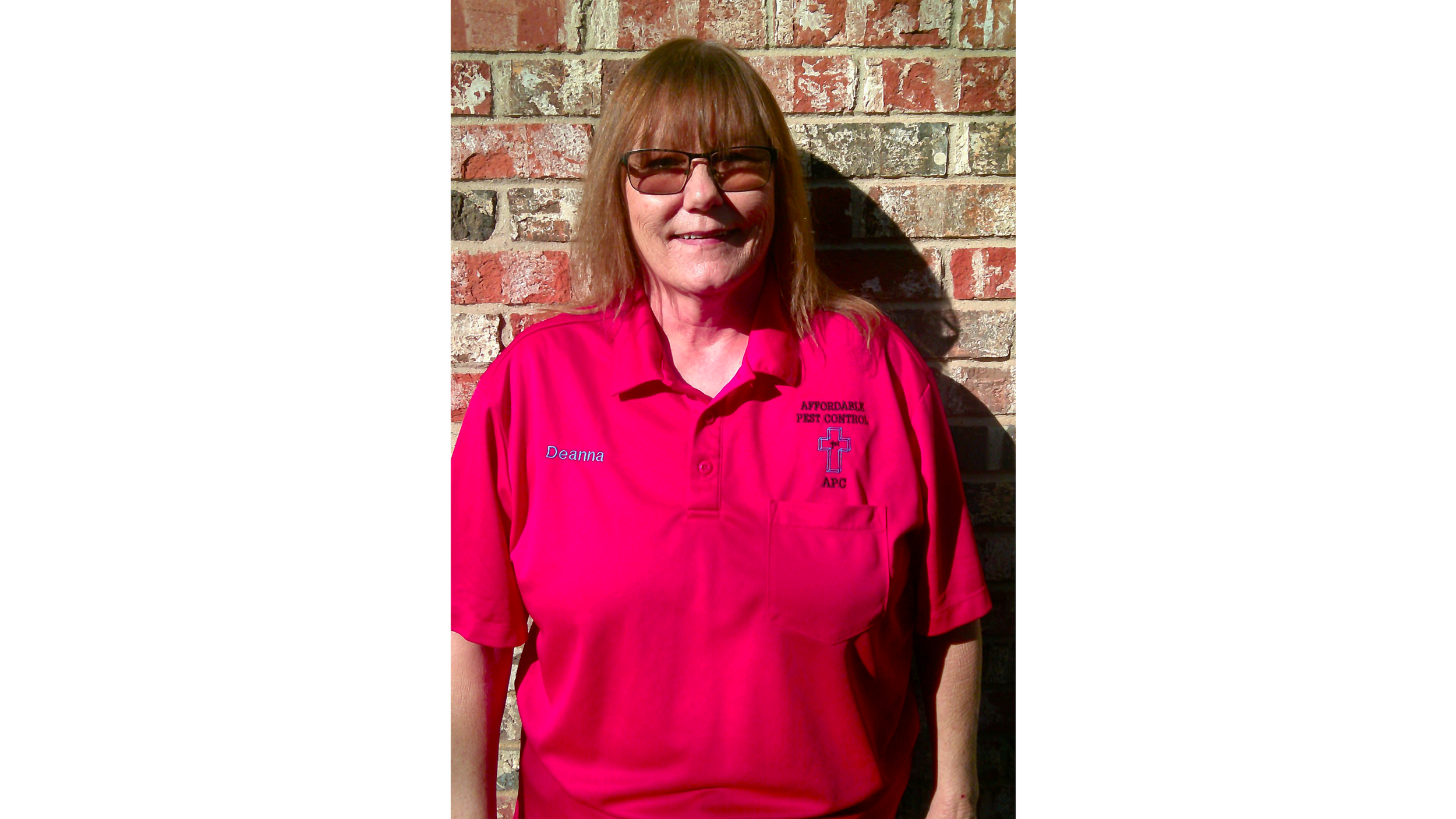A smiling woman with shoulder-length light brown hair and glasses, wearing a bright red polo shirt with the name tag 'Deanna' and the logo of 'Affordable Pest Control' in front of a brick wall.