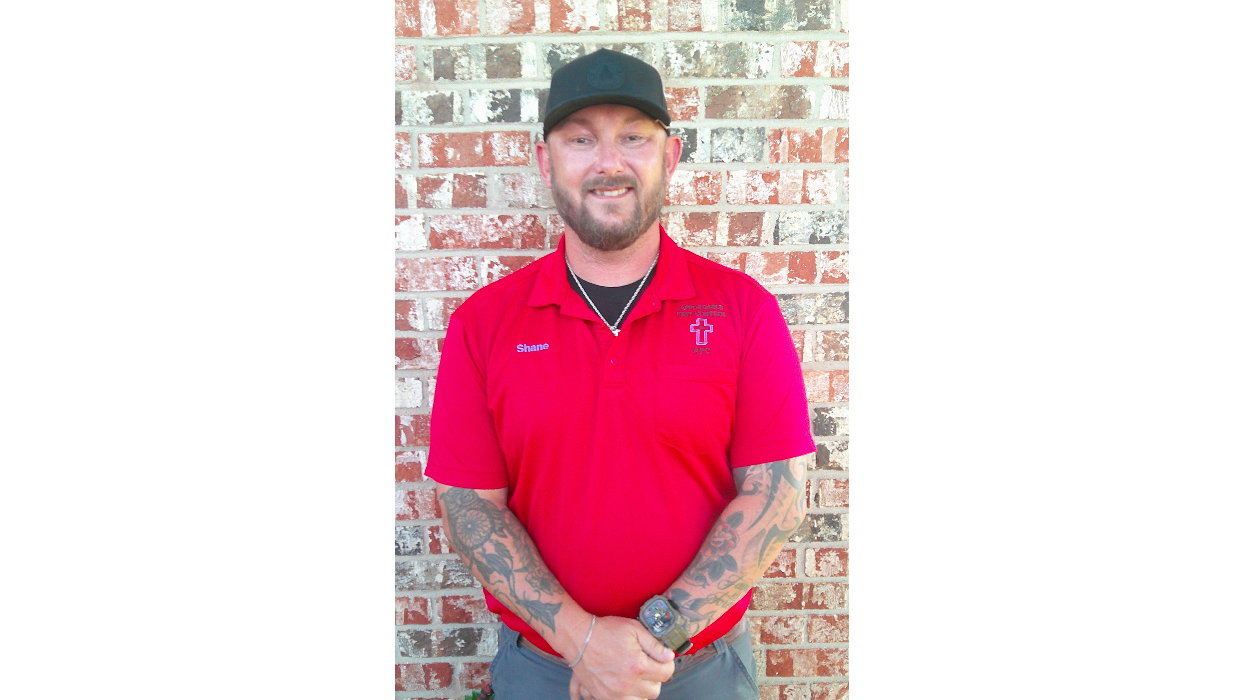 A man with tattoos, wearing a red shirt, glasses on his cap, and a watch, is sitting in an office talking on a landline phone, with multiple office supplies and papers on the desk, and a window with blinds behind him.