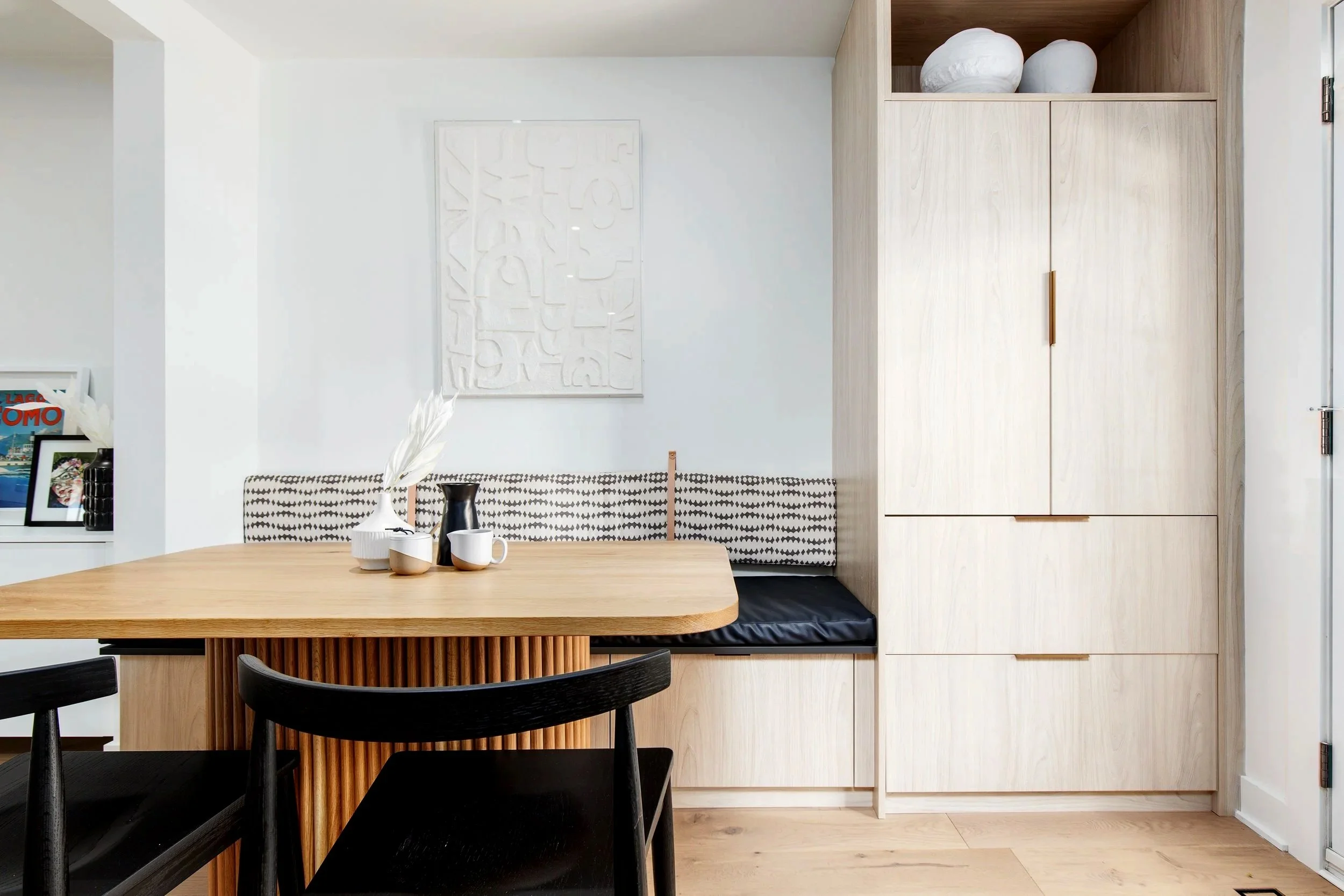 A modern dining area with a wooden table, black chairs, and built-in seating with black and white cushions against a white wall. There is a cabinet with decorative white pottery on the right side.