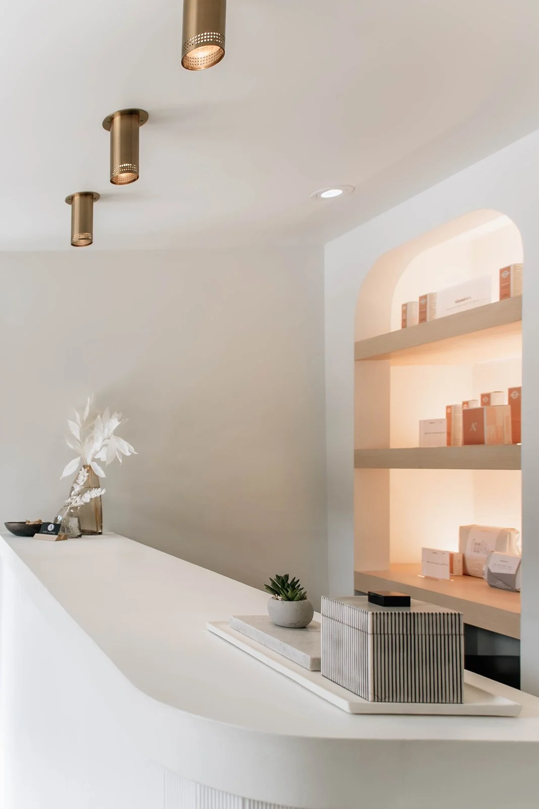 Minimalist reception area of a business with a white curved counter, small potted succulent, decorative striped box, and subtle golden ceiling lights.