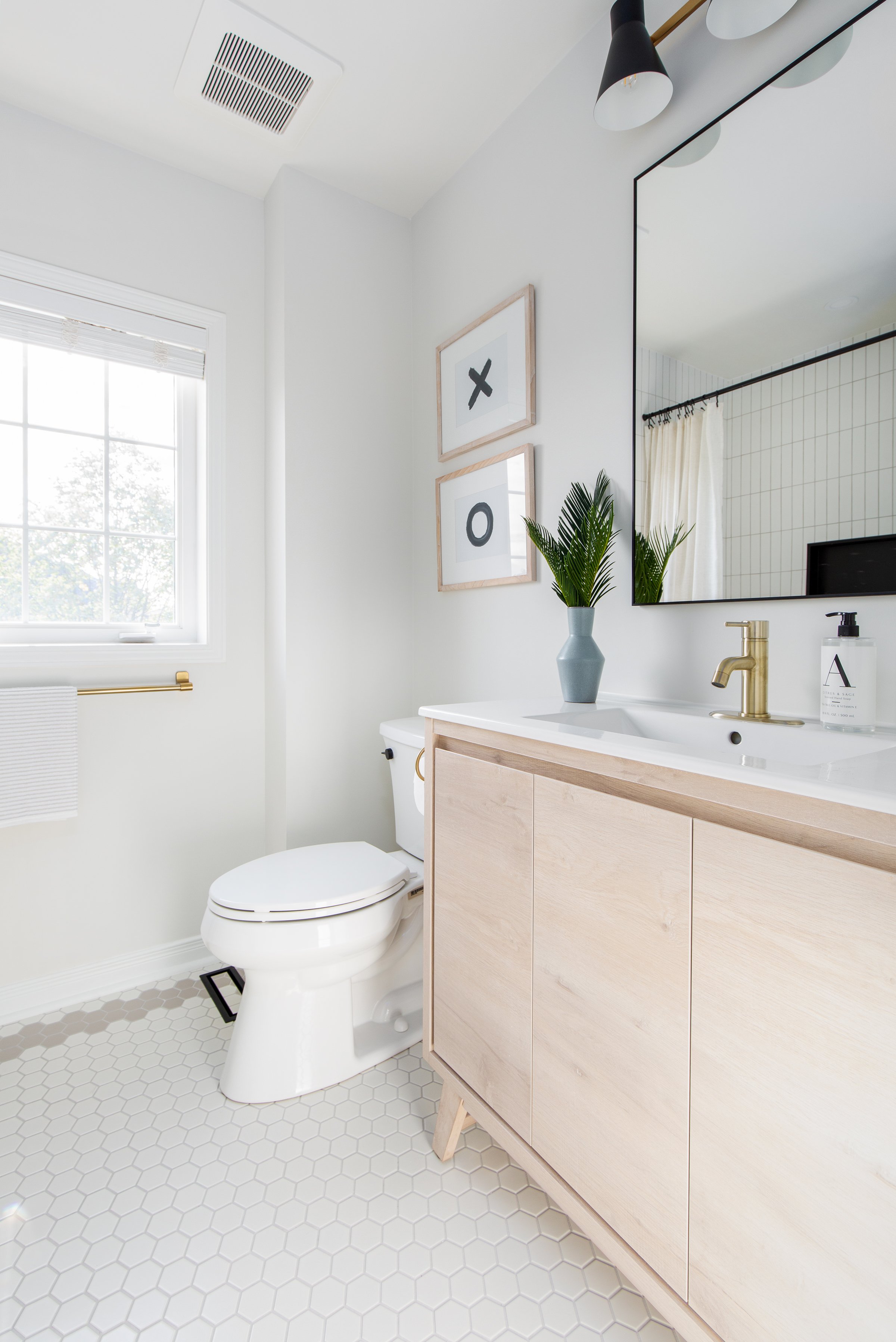 Modern bathroom with white hexagonal tile flooring, a white vanity with a wooden cabinet, a large mirror, and a toilet. Decor includes framed "X" and "O" art, a vase with green foliage, and a soap dispenser.