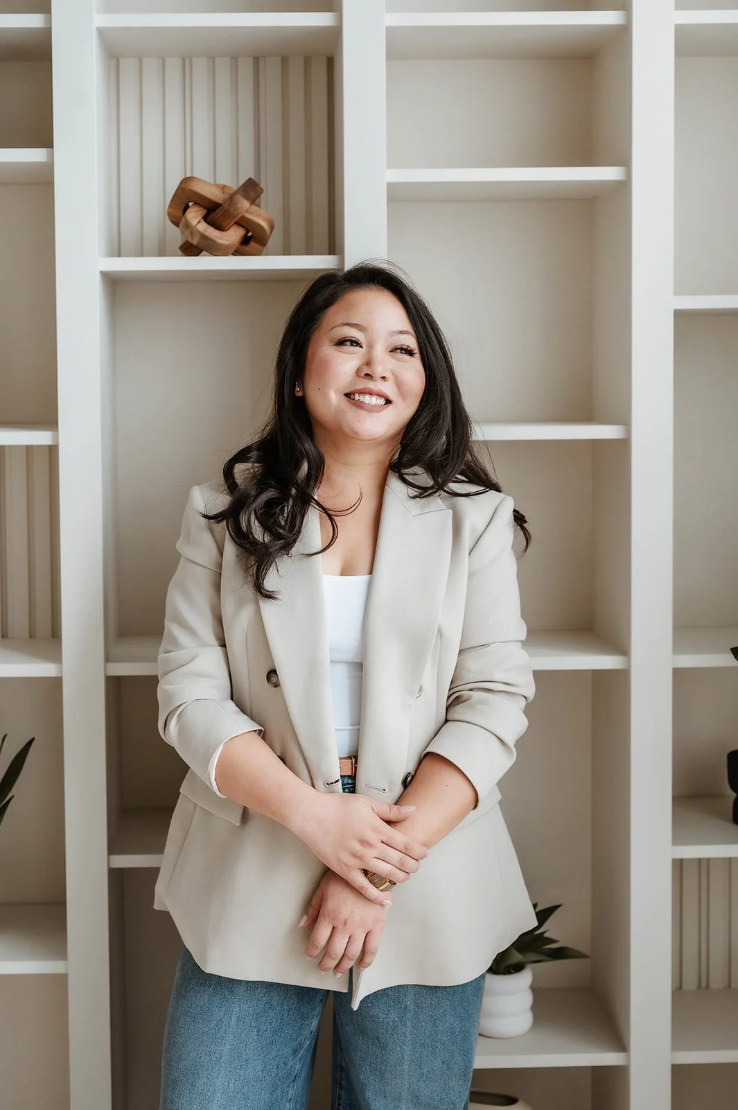Ruth Diana, the principal interior designer, with long, wavy black hair smiling, wearing a beige blazer and jeans, standing in front of a white bookshelf with decorative items and plants.