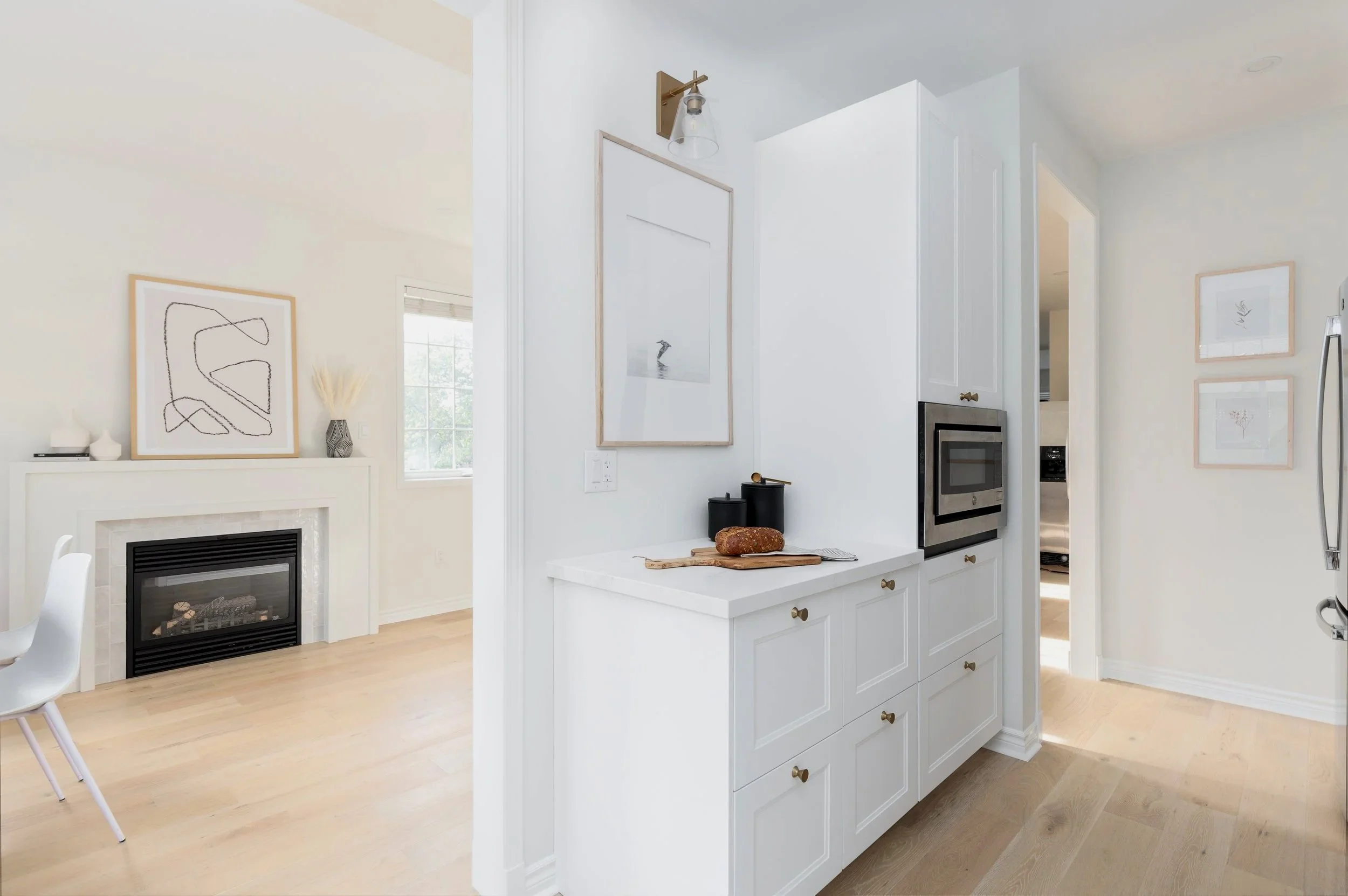 View of a modern home interior with a kitchen and living area. The kitchen has white cabinets, a built-in microwave, and some decorative items on the counter. The living area features a white fireplace with modern art and decor above it, and a window letting in natural light.