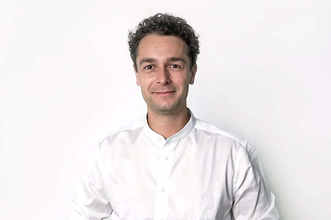 Ein Mann trägt ein weißes Hemd, hat dunkles, lockiges Haar und lächelt leicht.