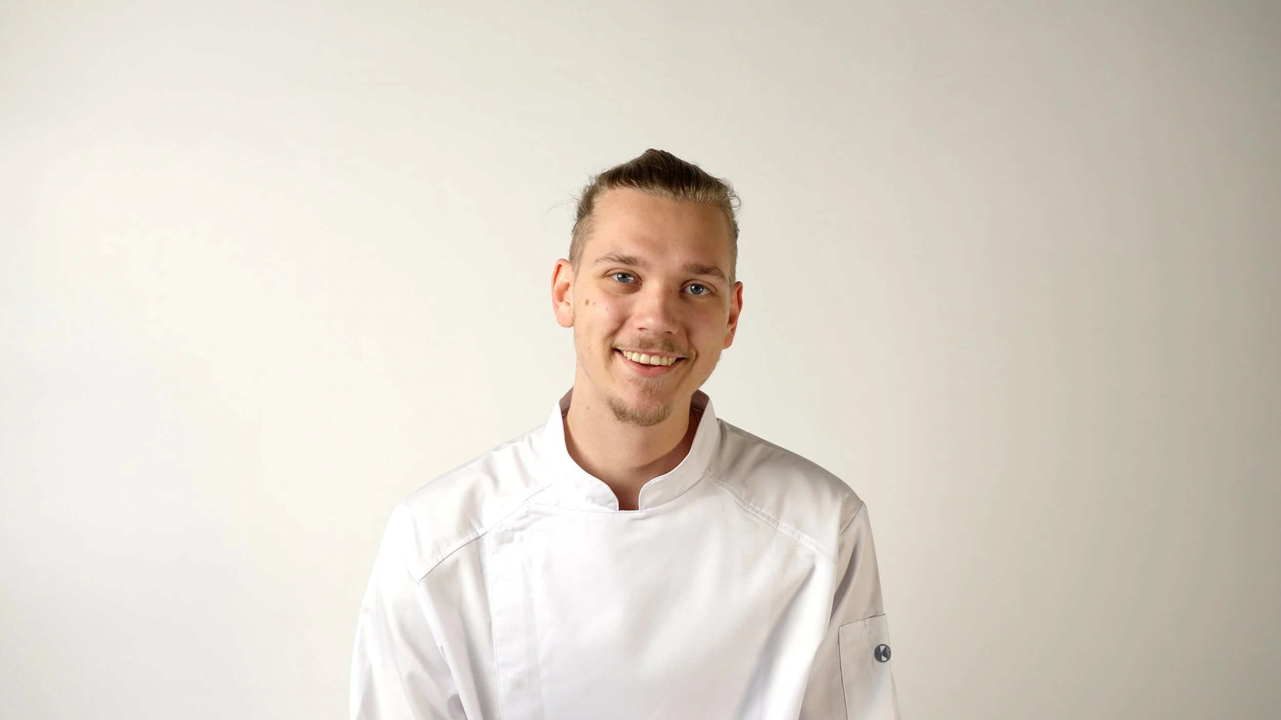 Ein junger Mann in einem weißen Kochkittel lächelt vor hellem Hintergrund.