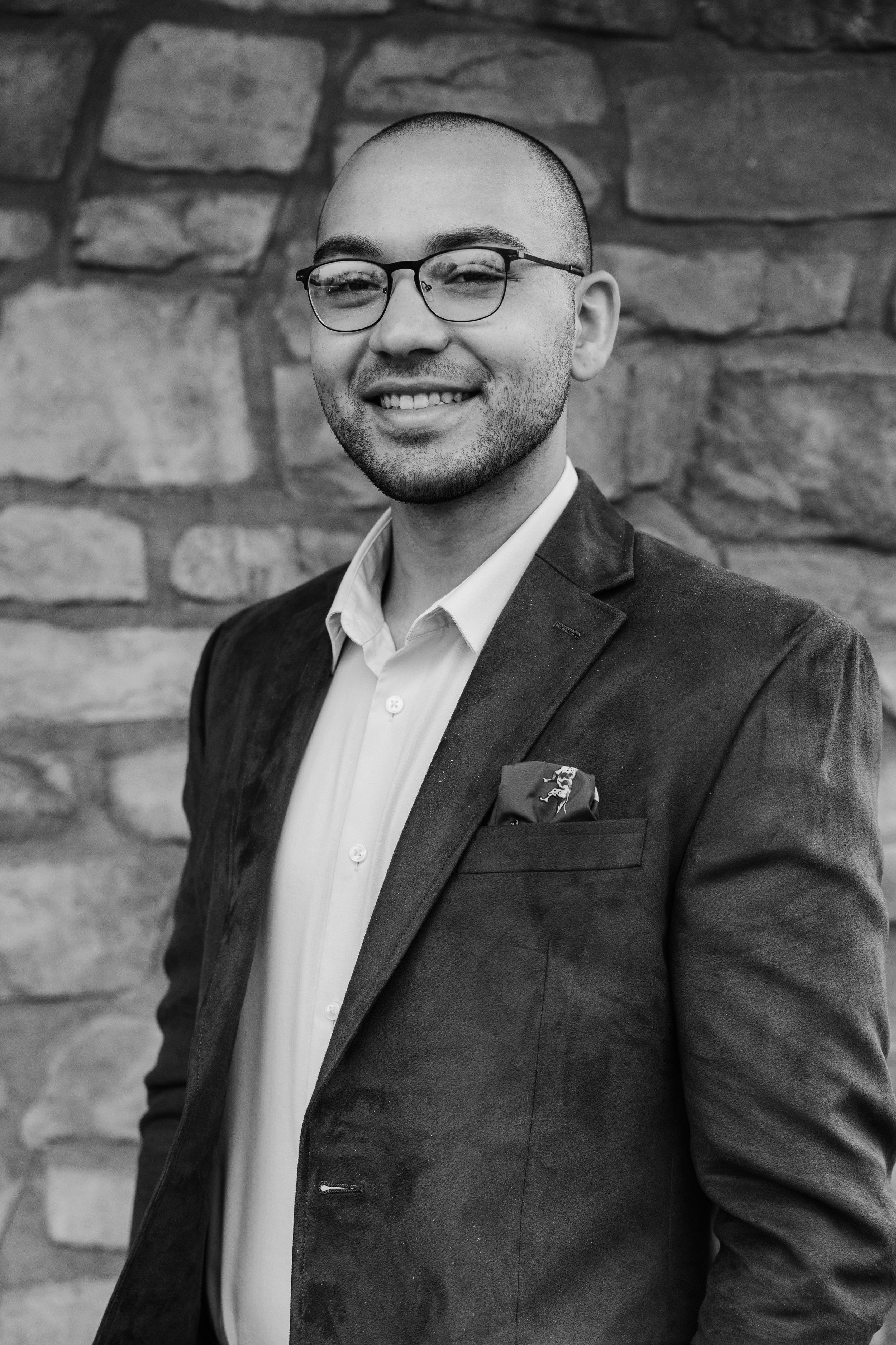 A smiling man with glasses, a groomed beard, dressed in a suit with an open collar shirt, and a pocket square, standing in front of a stone wall.